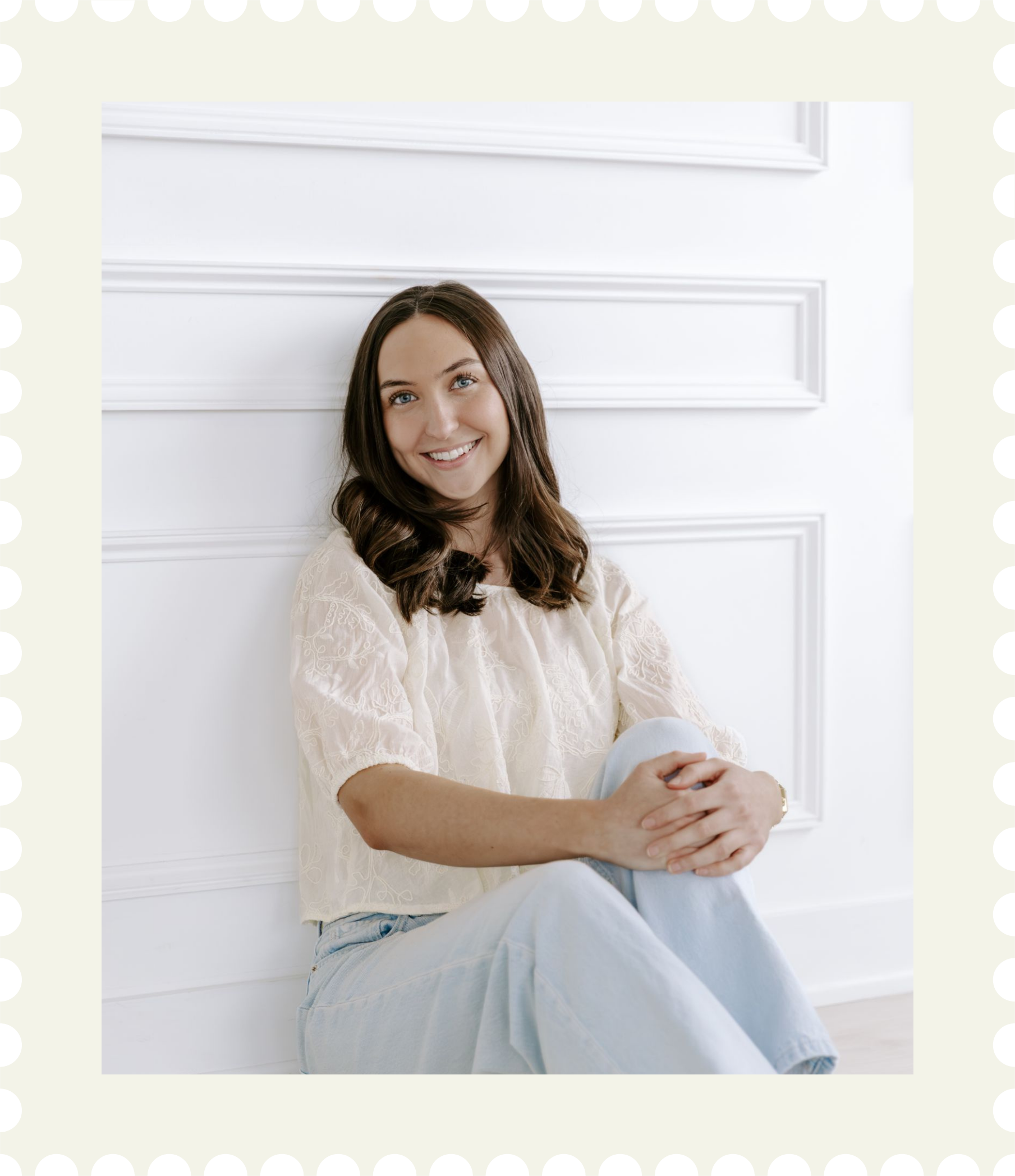 Young woman sitting cross-legged against a white paneled wall, smiling and looking at the camera for branding photography package in Ontario.