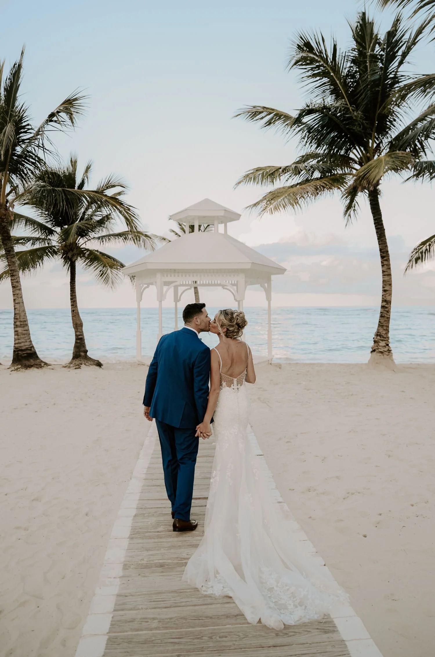 A newlywed couple kissing on a wooden pathway at the beach, surrounded by palm trees, with a white gazebo and ocean in the background for wedding photography package in Brockville Ontario.