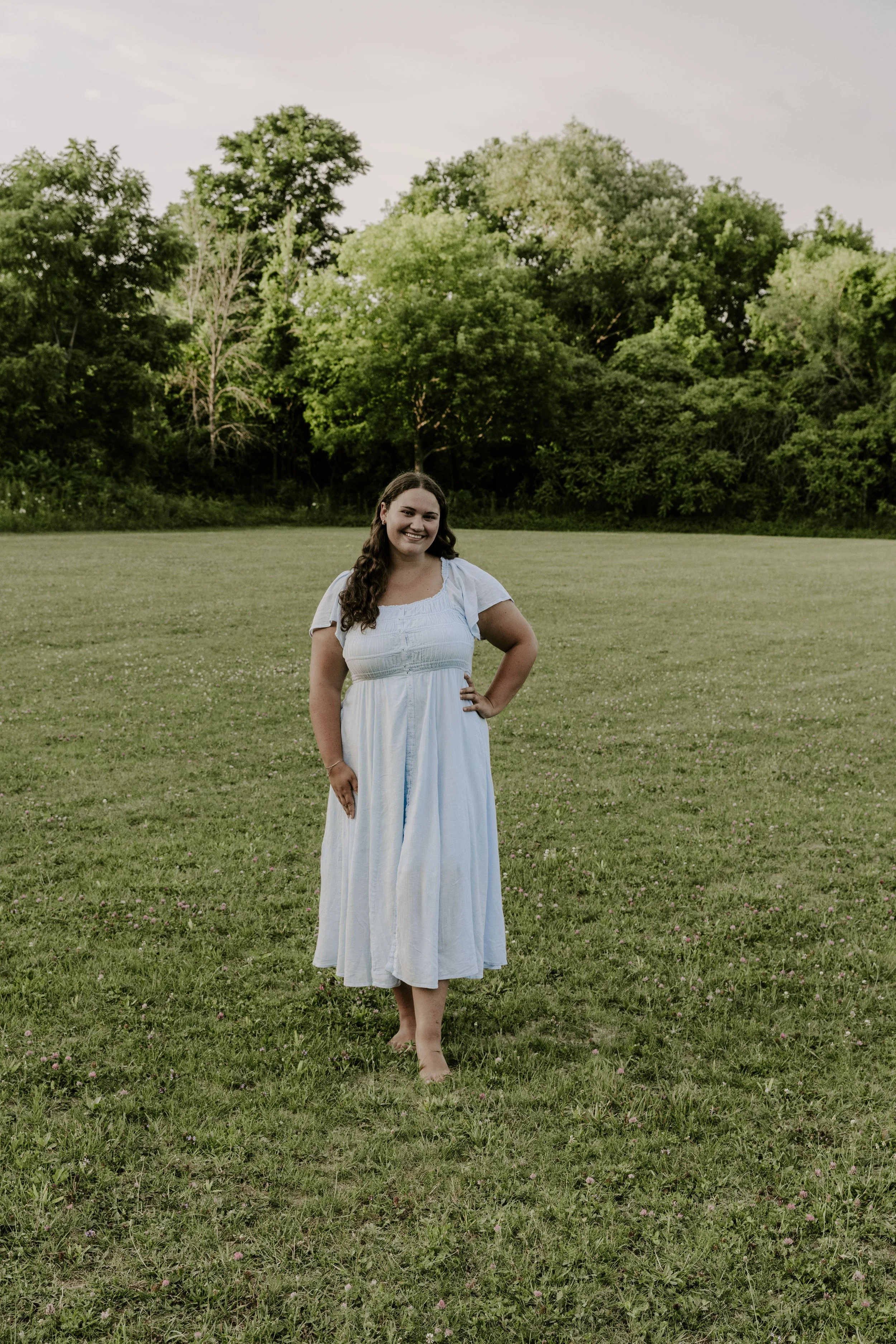A family, weddings, elopements and couples photographer standing barefoot on a grassy field with trees in the background, wearing a white dress and smiling.