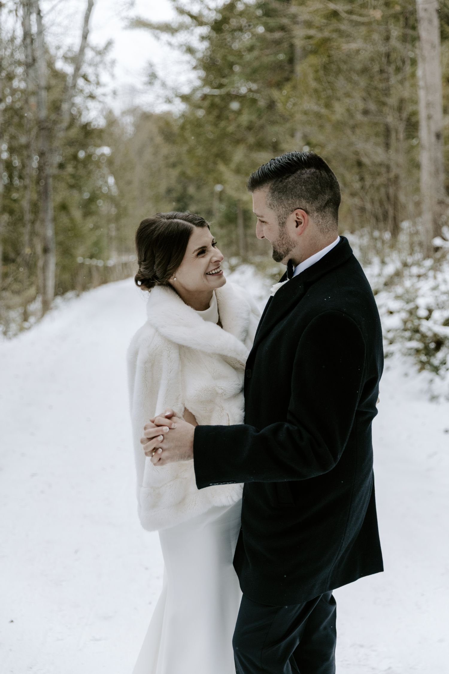 A bride and groom standing close together outdoors in a snowy forest, smiling and holding hands, sharing a loving moment during their wedding photography package in Ontario.