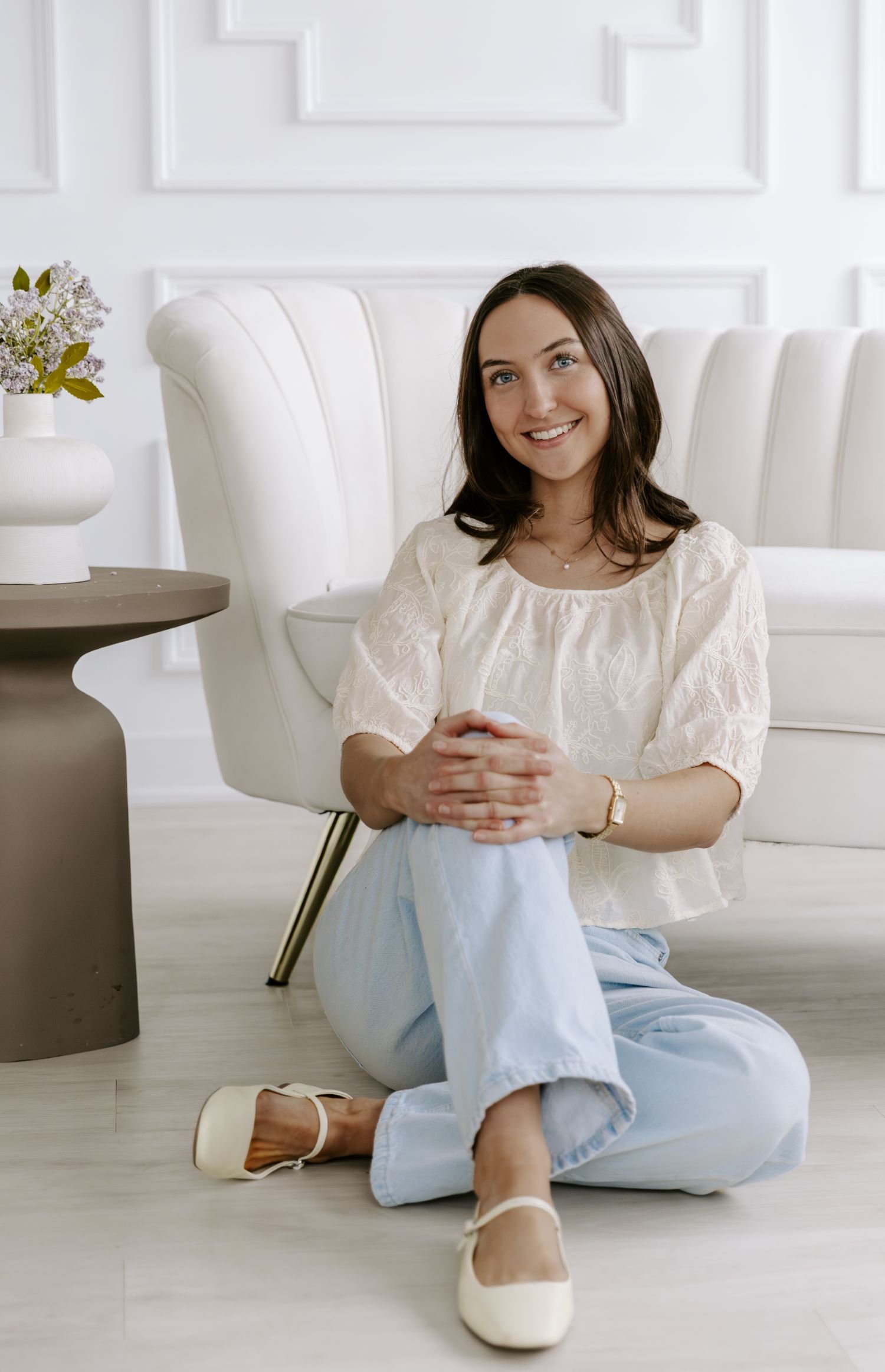 Young woman sitting on the floor with crossed legs, smiling, wearing a white blouse and light-colored pants, in a bright, modern living room with a white sofa and a side table with a vase of flowers during branding photography package in Brockville.