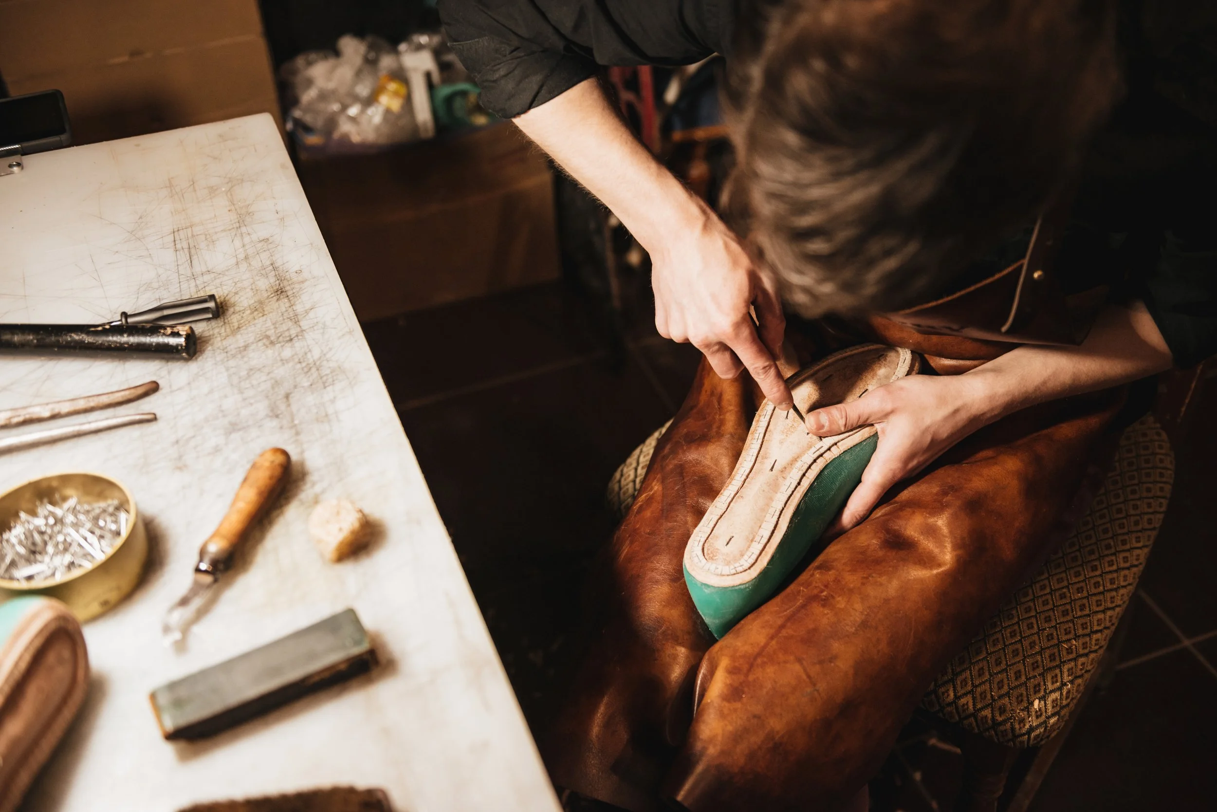 A cobbler crafts a shoe, carefully working on the sole with tools on a cluttered worktable in a workshop.