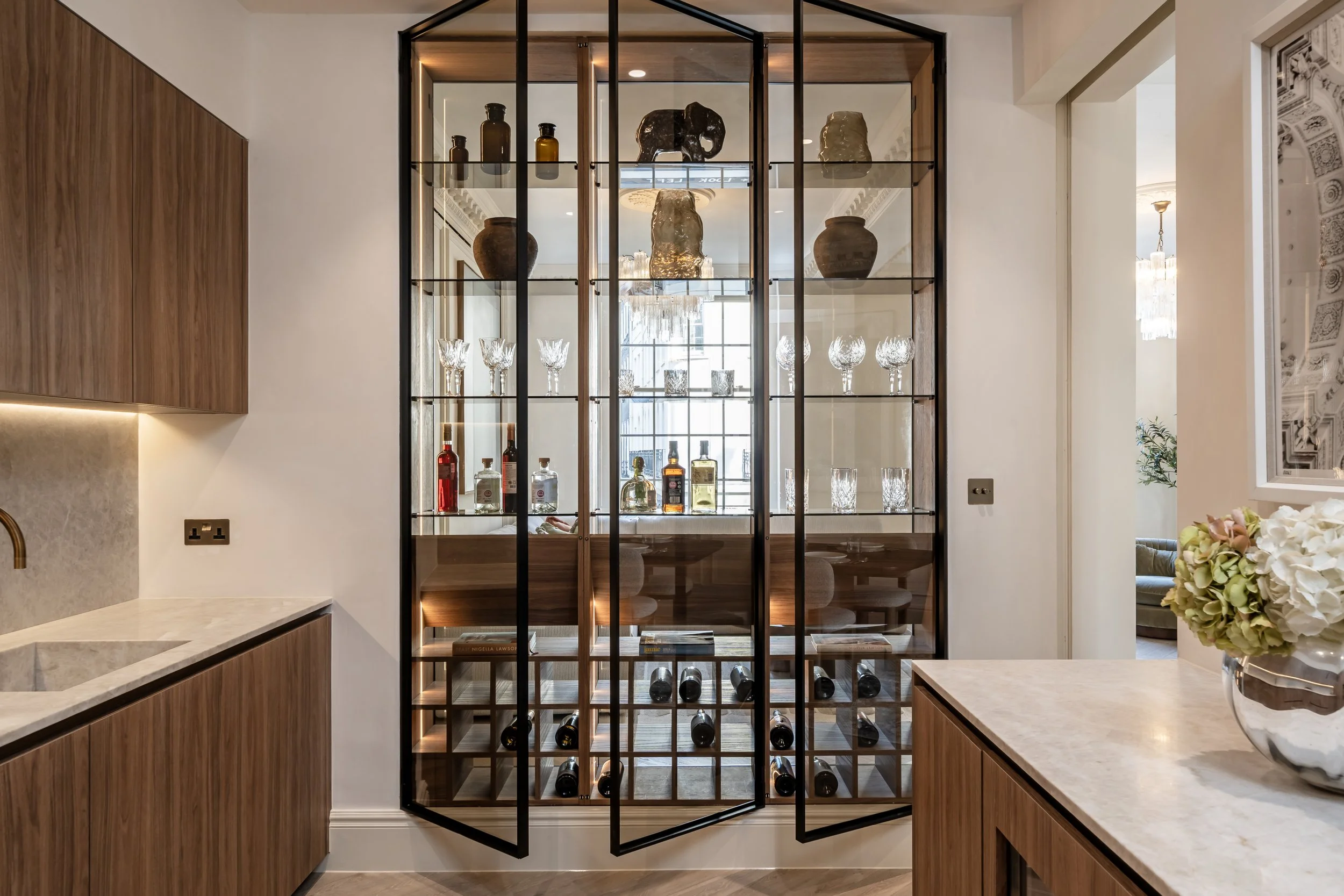A glass display cabinet filled with decorative bottles, glassware, and pottery, situated between two kitchen counters made of wood and marble in a modern home interior.