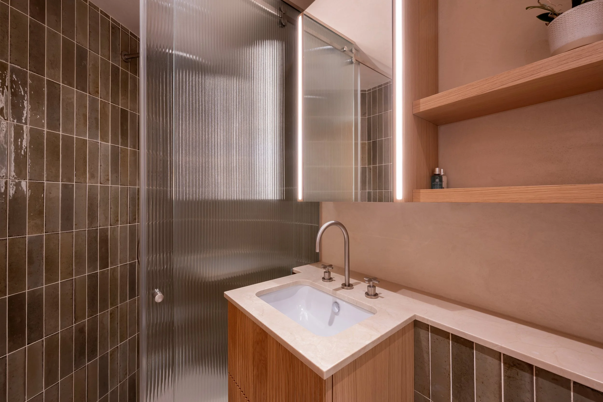 Bathroom with a small sink, a mirrored cabinet with lights, wooden shelves, and a partially visible shower enclosure with textured glass.