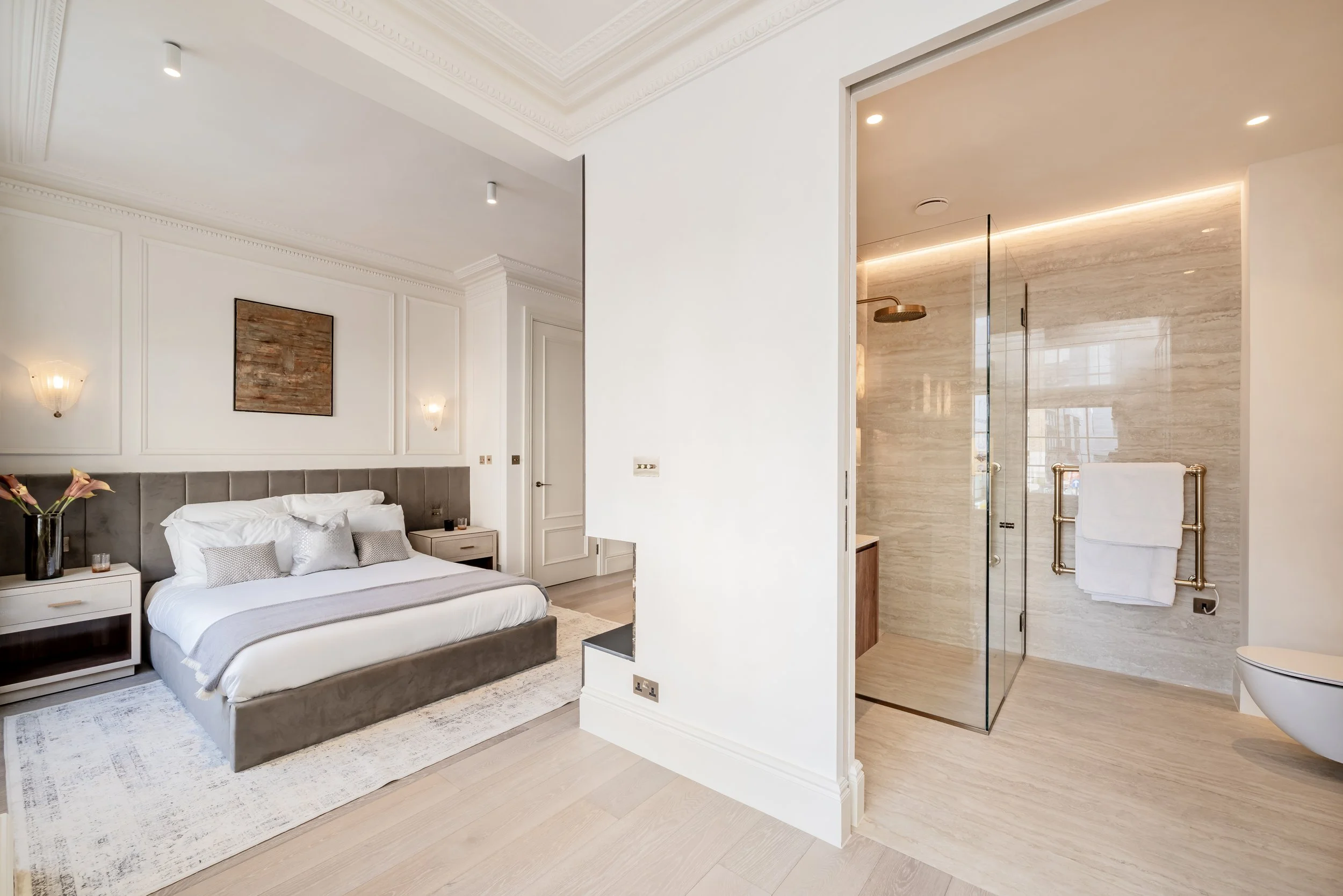 A luxurious bedroom with a beige and white color scheme, featuring a large bed with gray upholstery, bedside tables with lamps, and a framed artwork above the headboard. An open doorway reveals a modern glass shower with beige tiles and a heated towe