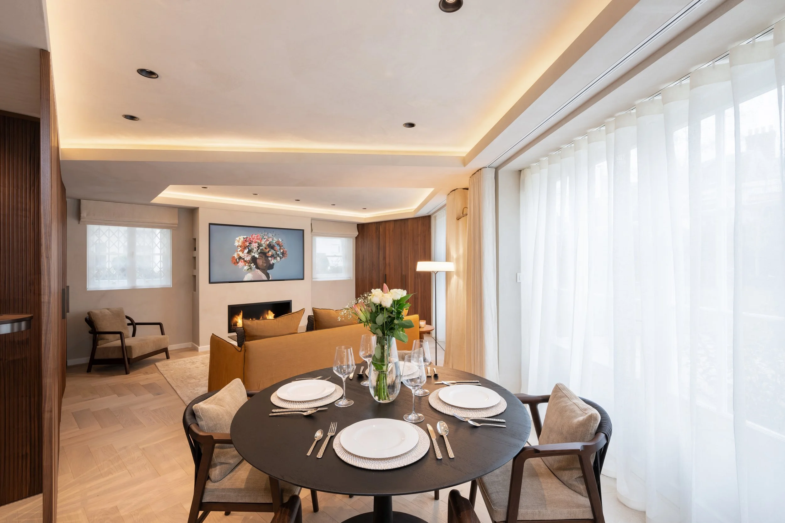 Dining room with a round table set for four, a flower vase, beige chairs, a sofa, a fireplace, a wall art, and large windows with sheer curtains.