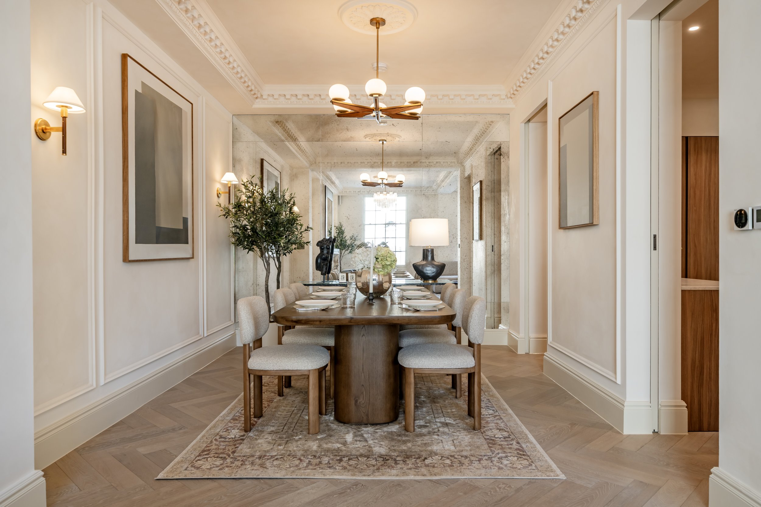 A dining room with a wooden table set with plates and silverware, surrounded by beige upholstered chairs, with a decorative rug underneath. The room has cream-colored walls with molding, brass wall sconces, and a large mirror on the back wall reflect