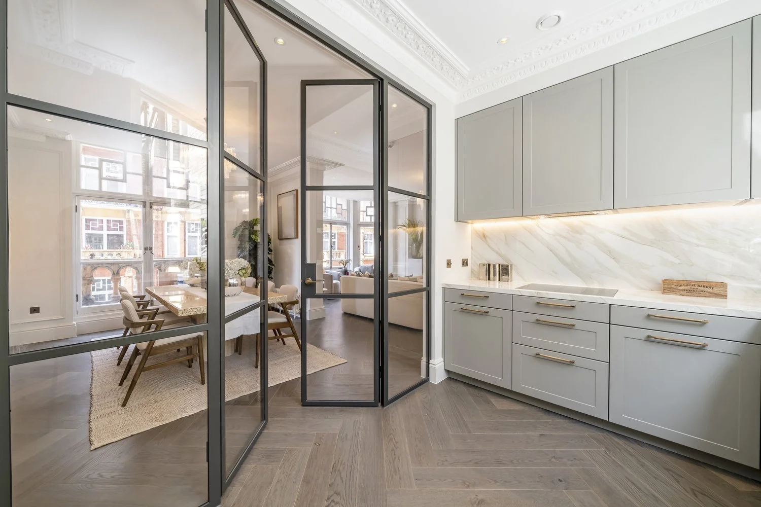 Modern kitchen with light gray cabinetry, marble backsplash, and wood flooring. Glass door separates kitchen from dining area, which has a large window, dining table, and chairs.