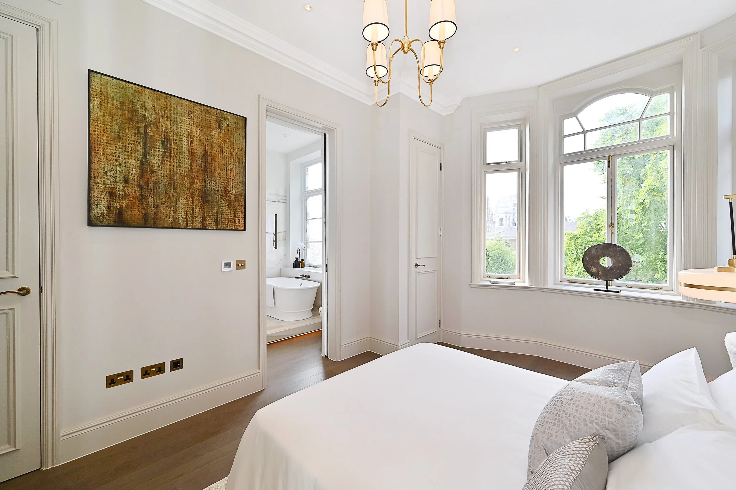 A bright bedroom with large bay windows, white walls, a modern chandelier, and a bed with white linens. An art piece hangs on the wall, and a view of a bathroom with a bathtub is visible through an open door.