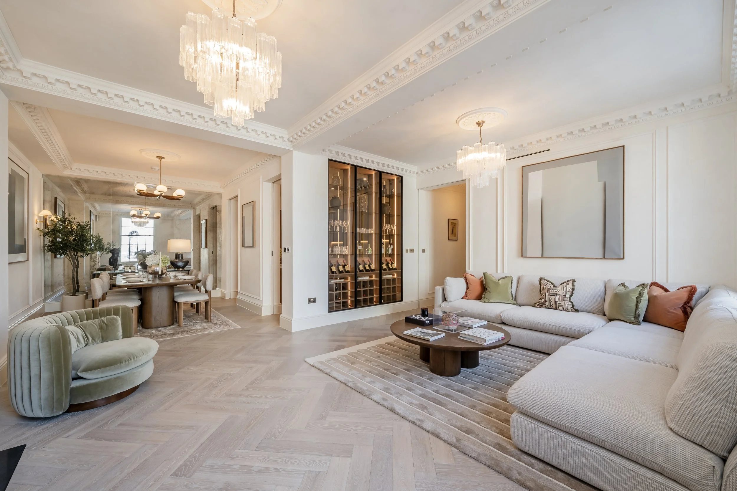 Elegant living room with white walls, colorful pillows on a long sectional sofa, a circular coffee table, a large mirror, and glass cabinet with glassware. Adjacent dining area with a large table and multiple chairs, chandelier lighting, and potted p