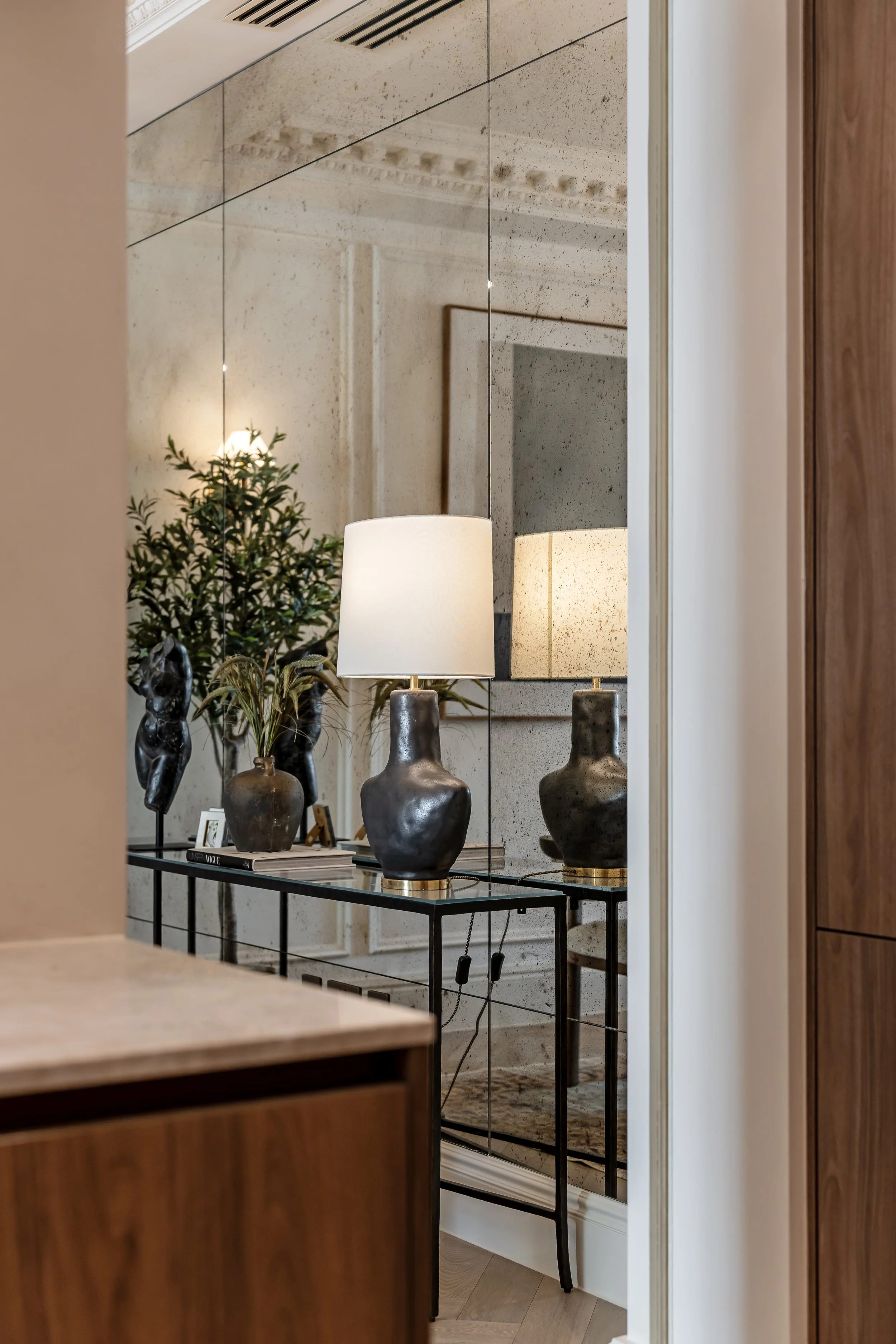 Interior view of a modern hallway with a table holding two lamps and decorative vases, reflected in a large mirror on the wall.