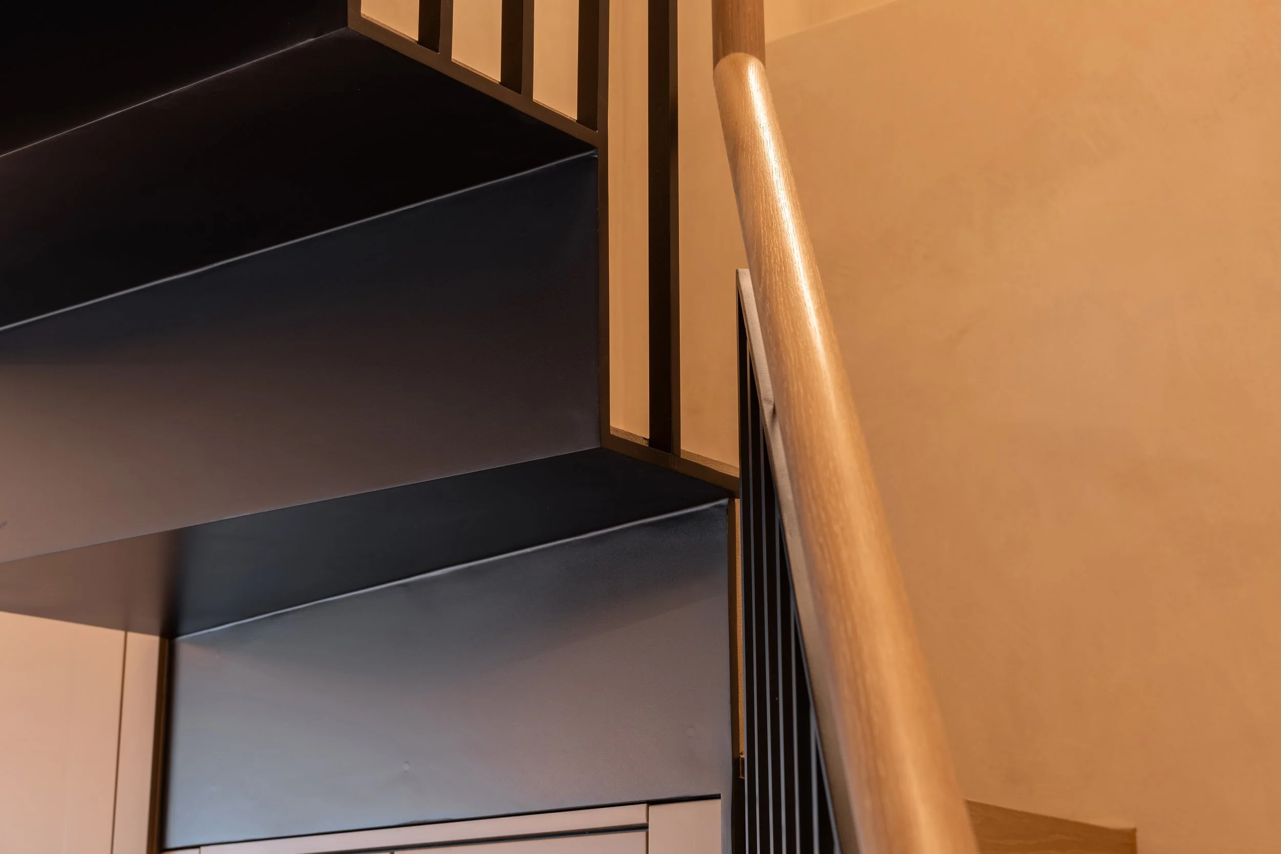 A staircase with a wooden handrail and black metal balusters, next to a beige wall, with the underside of a black and beige staircase visible.
