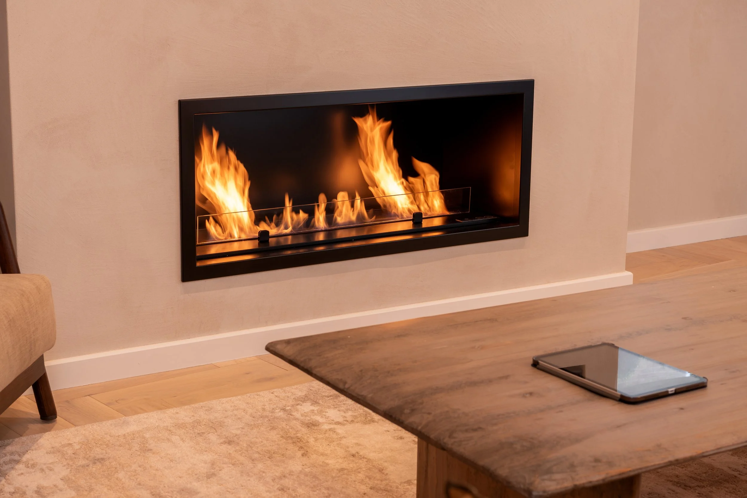 Modern living room with a wall-mounted fireplace with visible flames, a wooden table with a tablet on it, a beige carpet, and a beige wall.