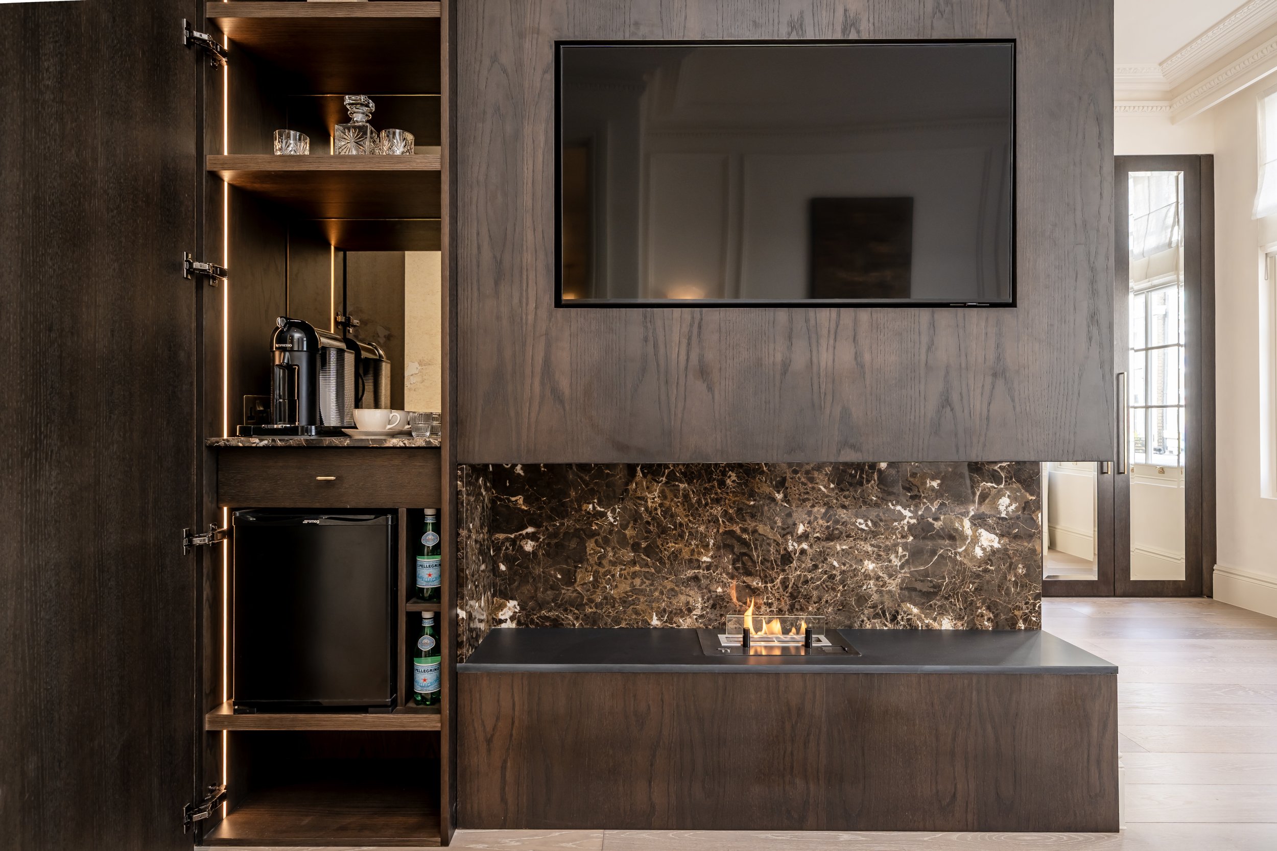 Interior of a modern living room with a wood-paneled wall, a flat-screen TV, a built-in shelf with glassware, coffee machine, mini fridge, and a fireplace with a marble surround.