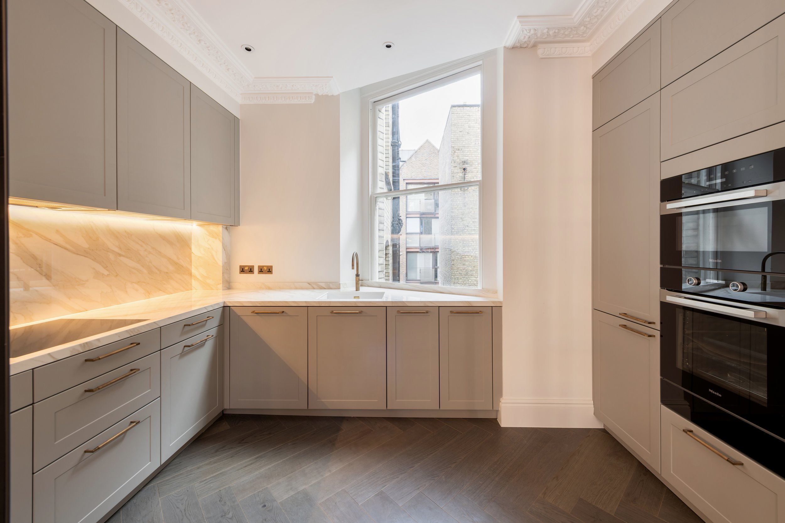 Kitchen with gray cabinets, marble countertops, a stainless steel sink with a brass faucet, and a large window overlooking neighboring buildings.