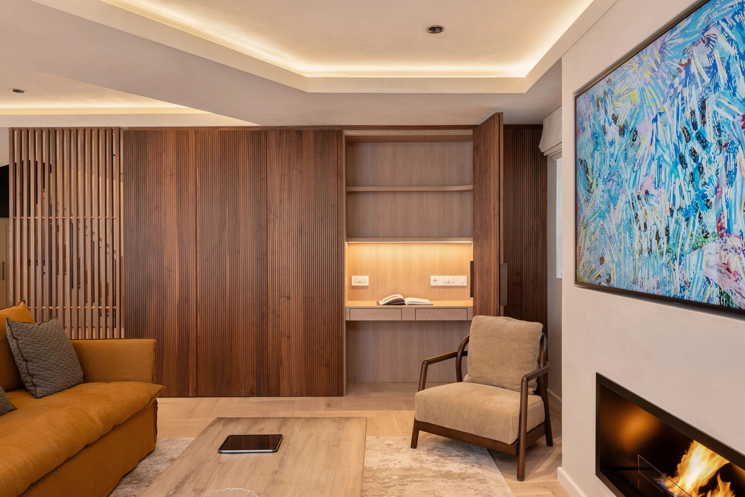 Interior of a modern living room with a wooden accent wall, beige armchair, mustard-colored sofa, large colorful abstract painting, and a fireplace with visible flames.