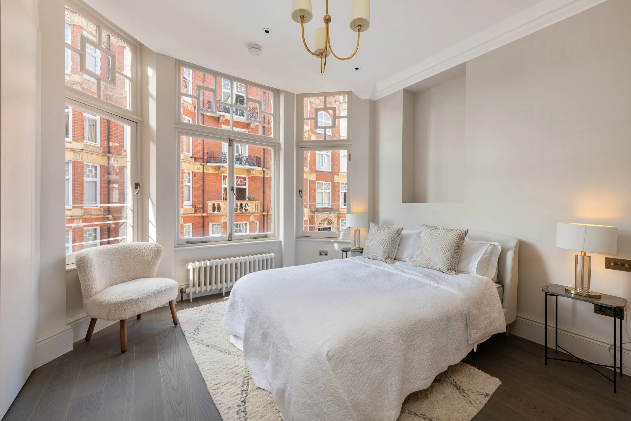 Bright bedroom with large windows, white bedding, a cream armchair, and modern lamps.