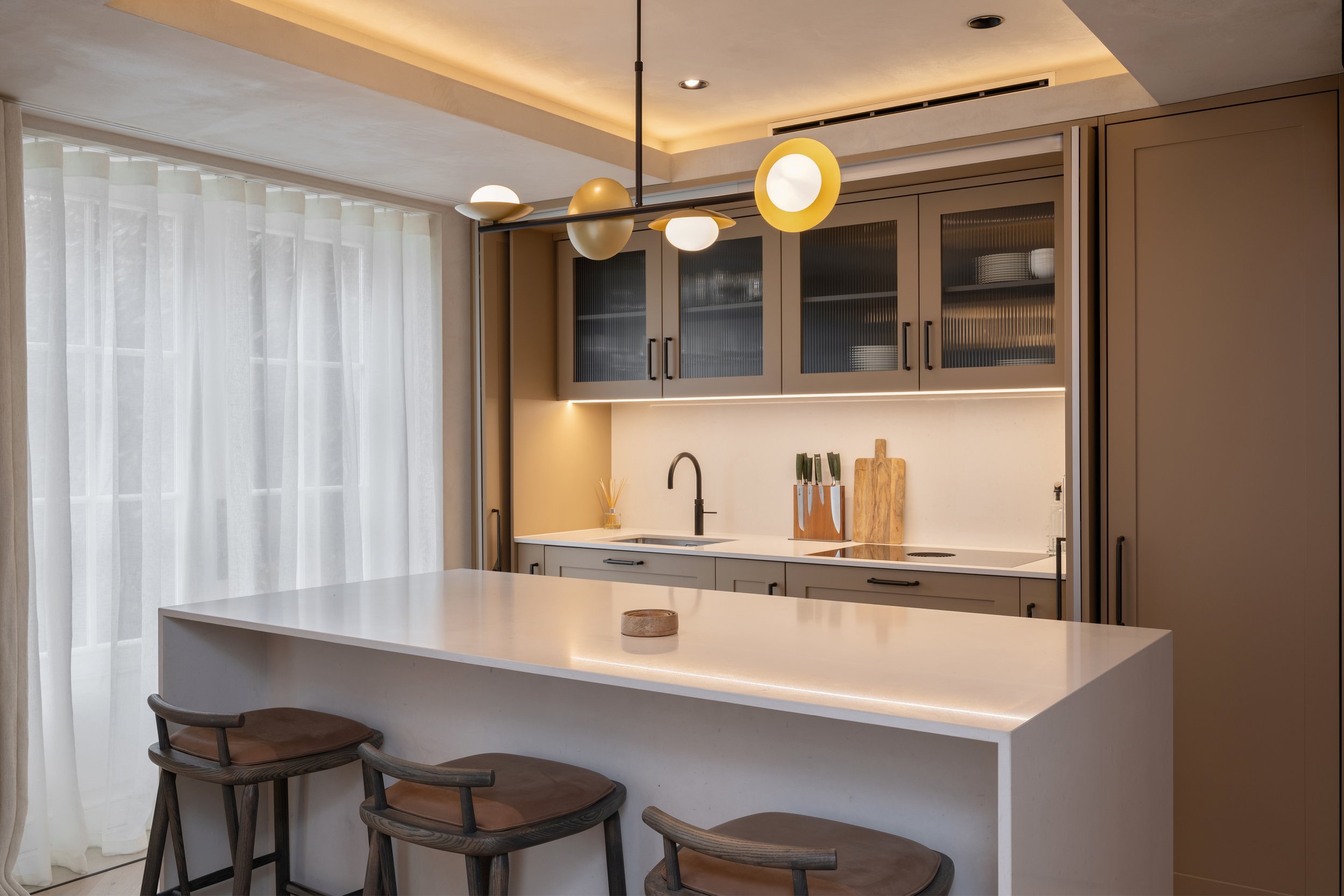 Modern kitchen with a white island, three bar stools, beige cabinetry, a black faucet, and a decorative light fixture.