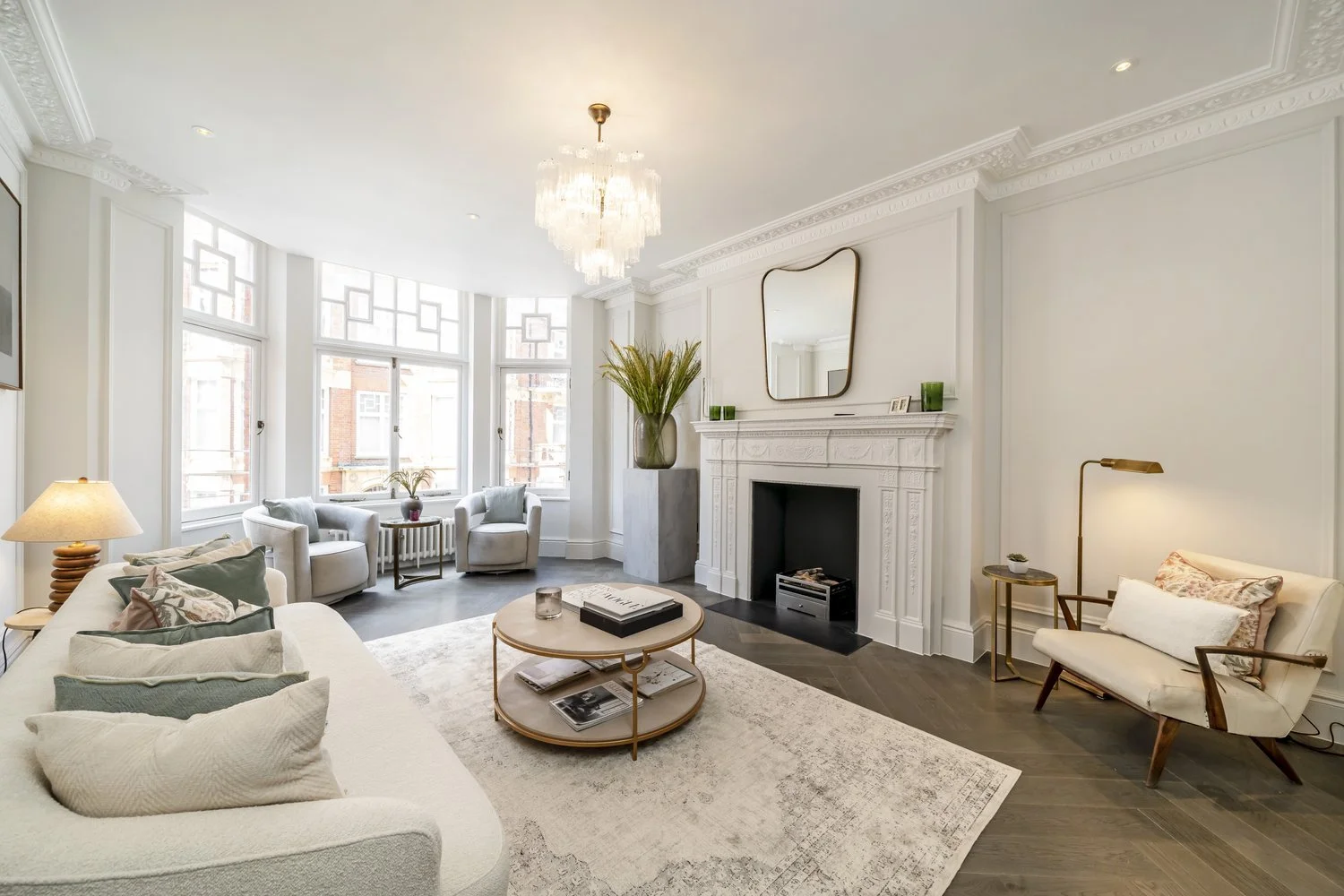 Bright living room with white walls, decorative molding, a white fireplace, a large mirror above the fireplace, a chandelier, large bay windows, a cream sofa with pillows, a round coffee table with books and candles, a cushioned armchair with a side 