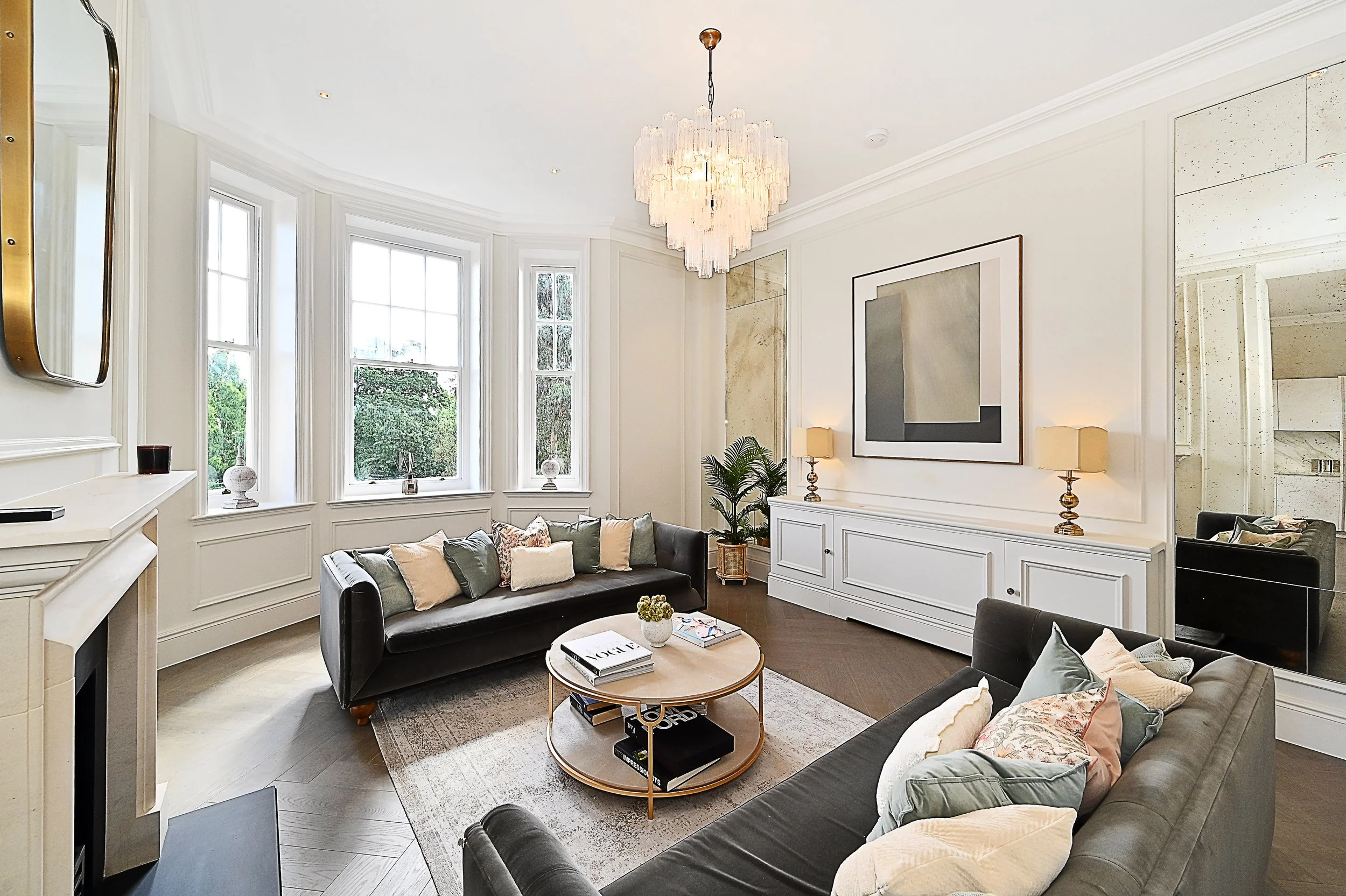 Living room with white walls, large windows, black sofas, a round coffee table with books and a plant, modern art on the wall, and a chandelier hanging from the ceiling.