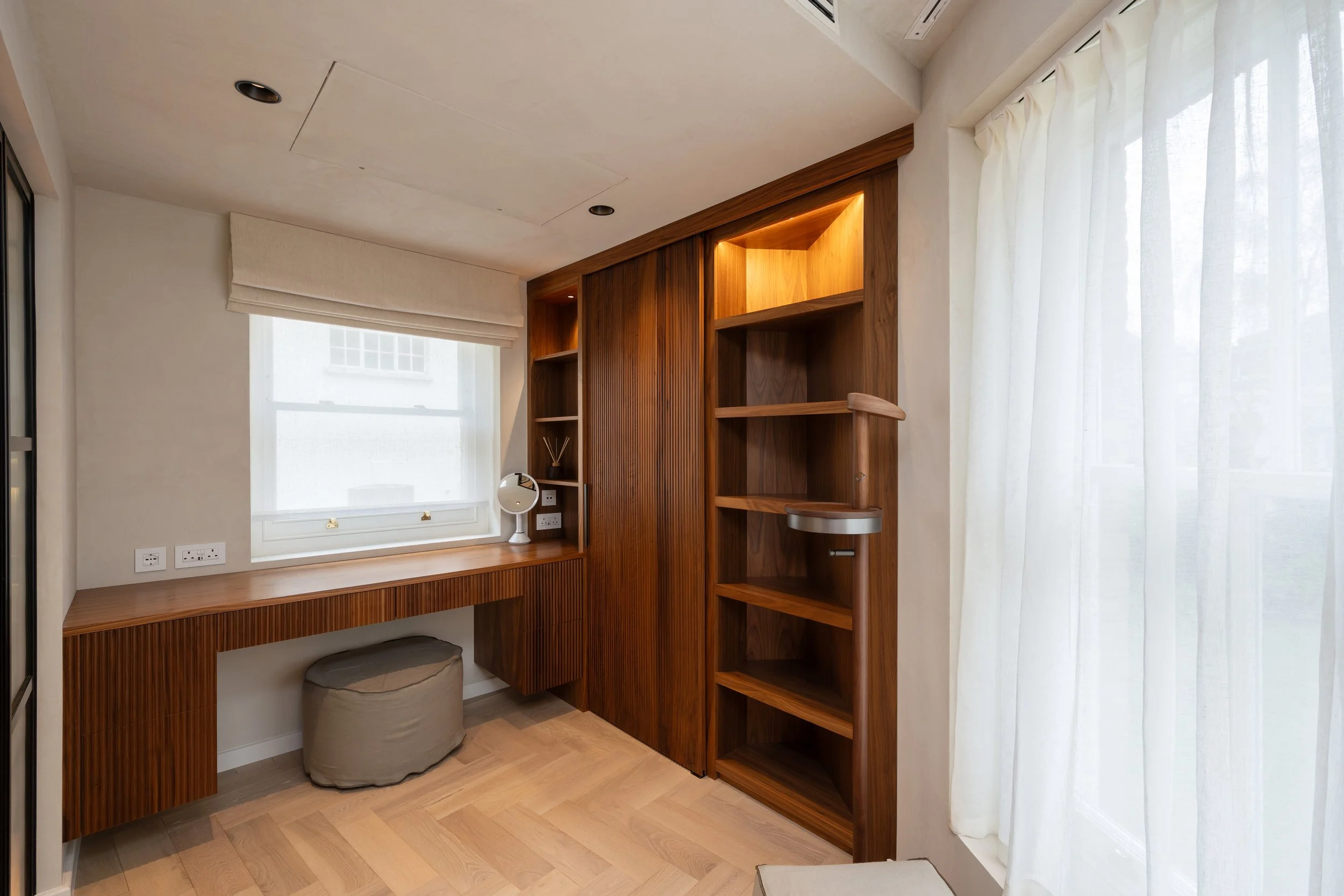 Home office with a built-in wooden desk and shelves, a small round mirror, and a beige ottoman near the window, with light-colored curtains and wood flooring.