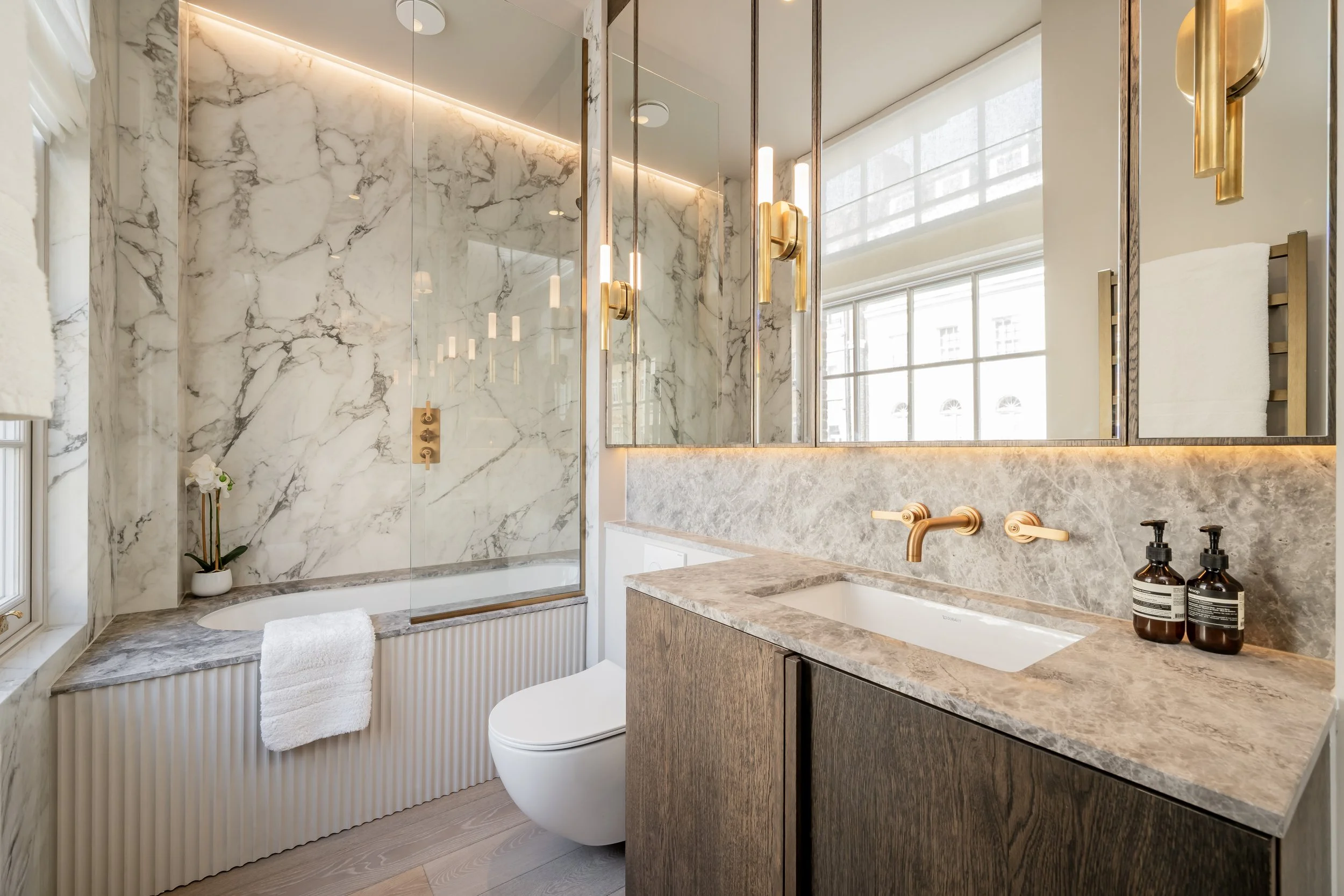 Modern bathroom with marble walls, a bathtub, a toilet, a sink with a marble countertop, large mirror, and gold fixtures, illuminated by natural light from a large window.