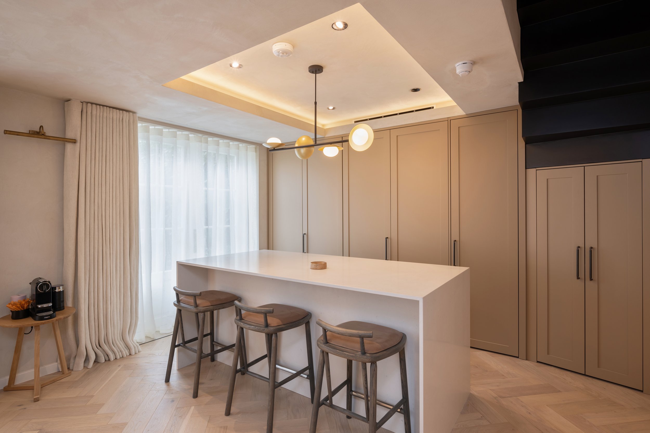 Modern kitchen with beige cabinets, a white island, and three bar stools. There is a window with sheer curtains, a small side table with a coffee machine, and a contemporary ceiling light fixture.