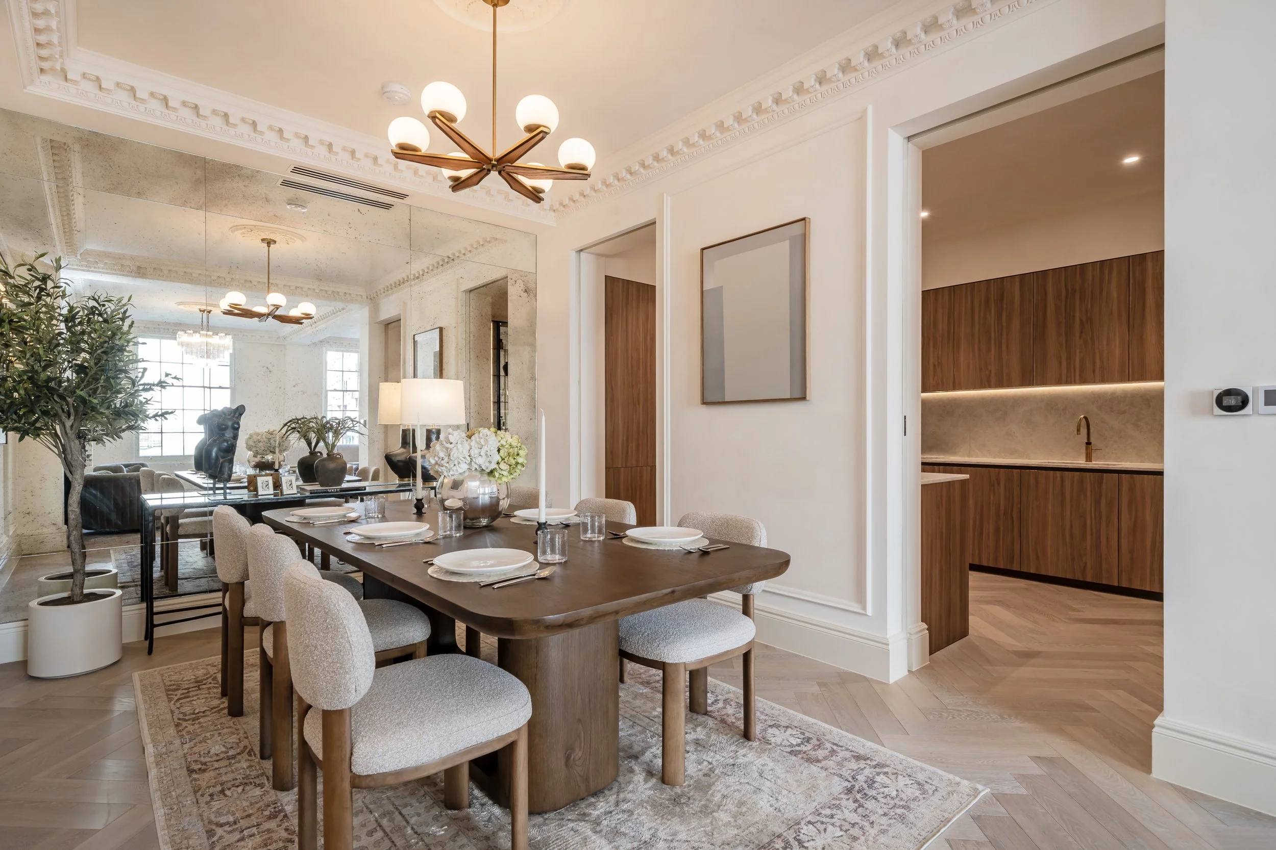 Dining room with a wooden table set for six, surrounded by light beige chairs, with a floral centerpiece and candles. A large mirror reflects part of the room, and there are modern light fixtures and a green potted plant. A doorway leads to a kitchen