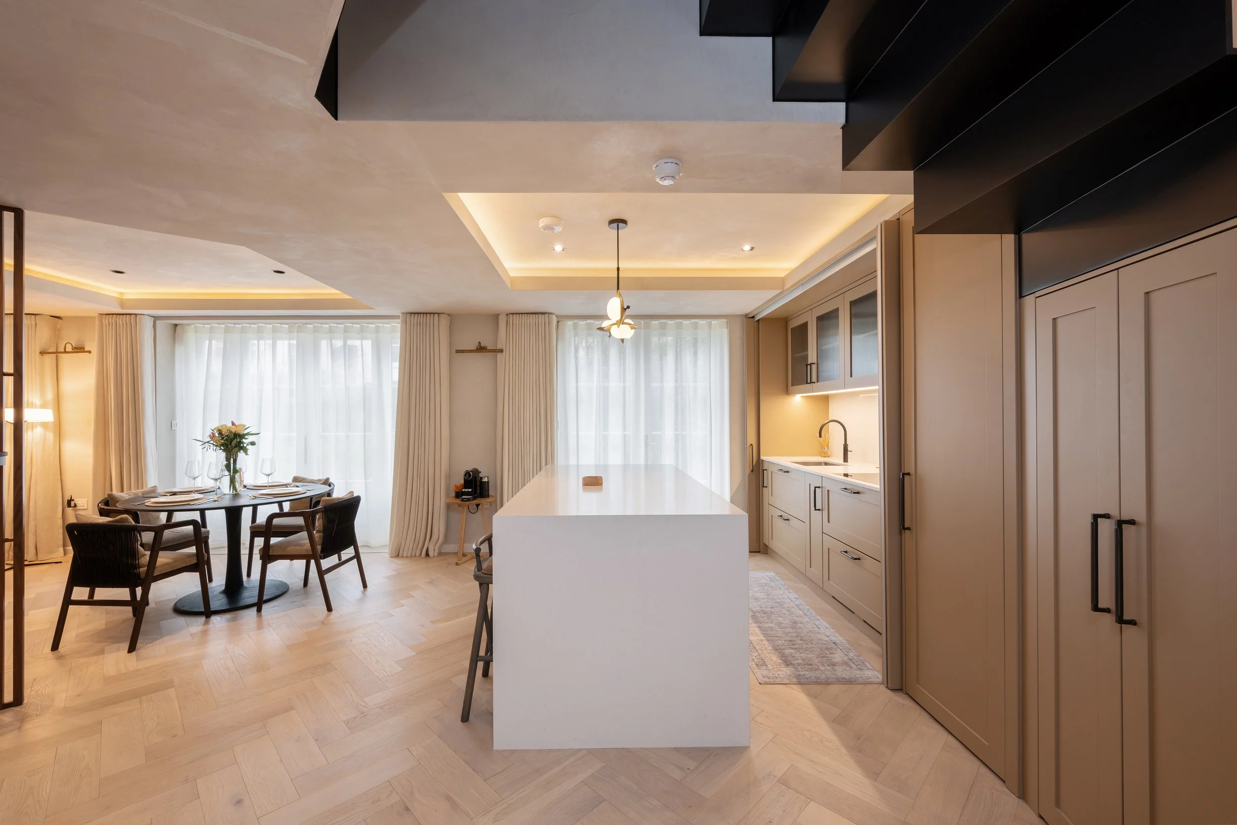 Open-concept kitchen and dining area with beige cabinetry, white countertops, and light wood flooring. A round dining table with four chairs and a floral centerpiece is near large windows with sheer curtains, while the kitchen features an island, han