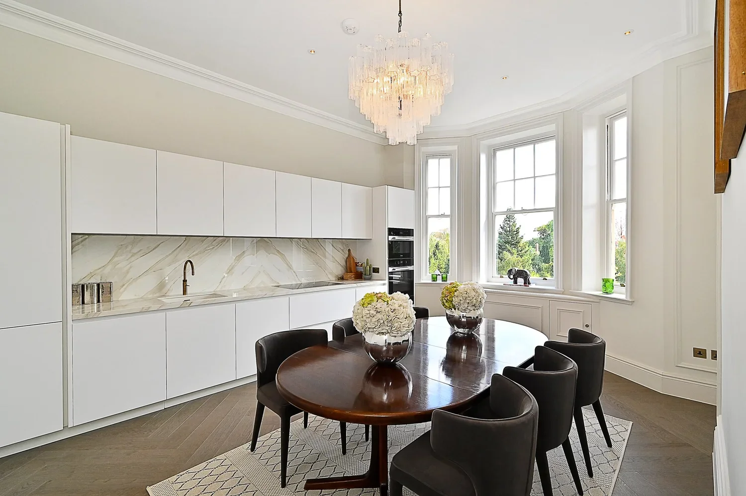 Bright modern dining room with large bay windows, white cabinetry, marble backsplash, dark wood oval table with black chairs, and floral centerpieces, chandelier, and decorative items.