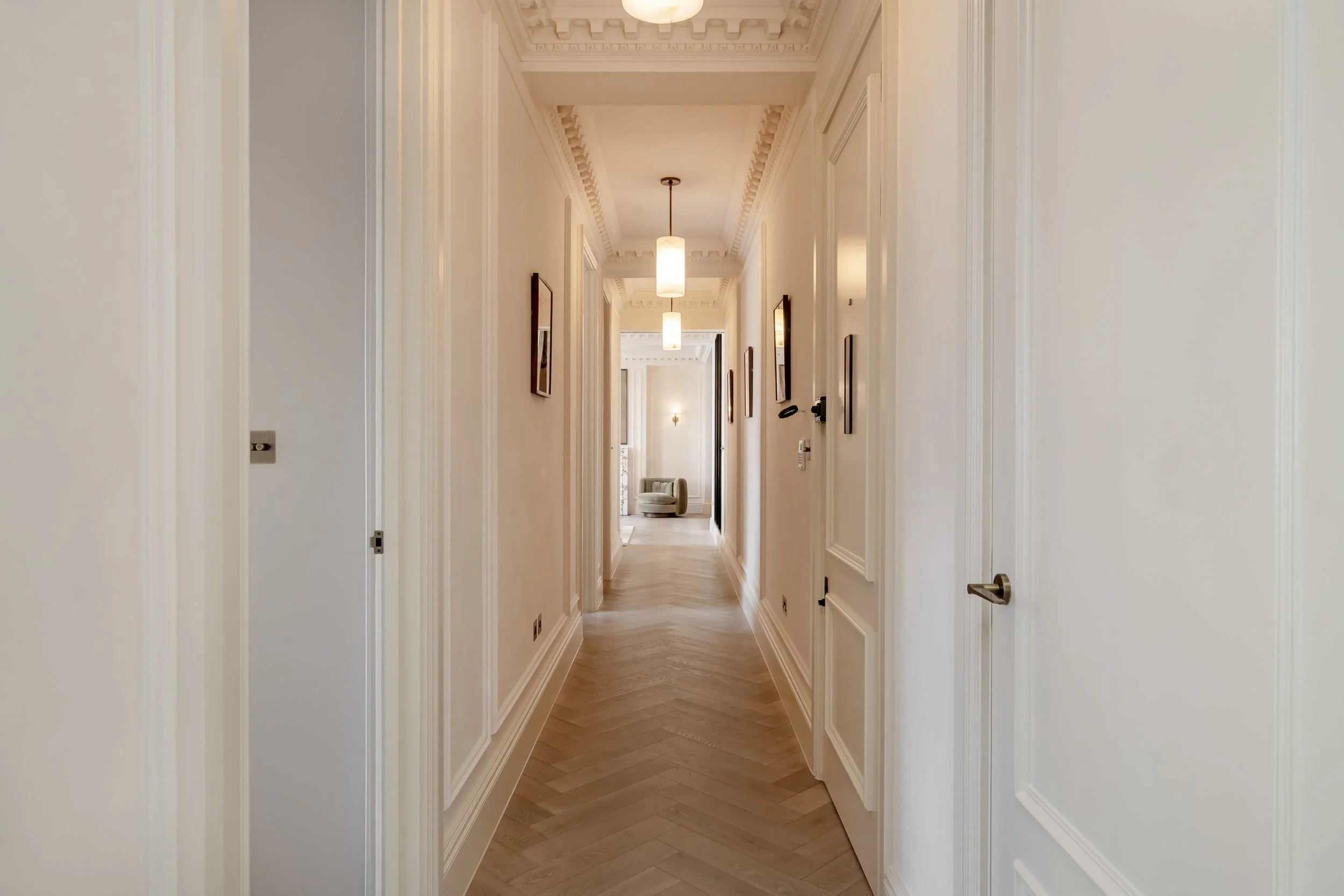 Elegant hallway with white walls, decorative crown molding, hanging pendant lights, and a light wood herringbone floor, leading to a sitting area with a green armchair at the end.