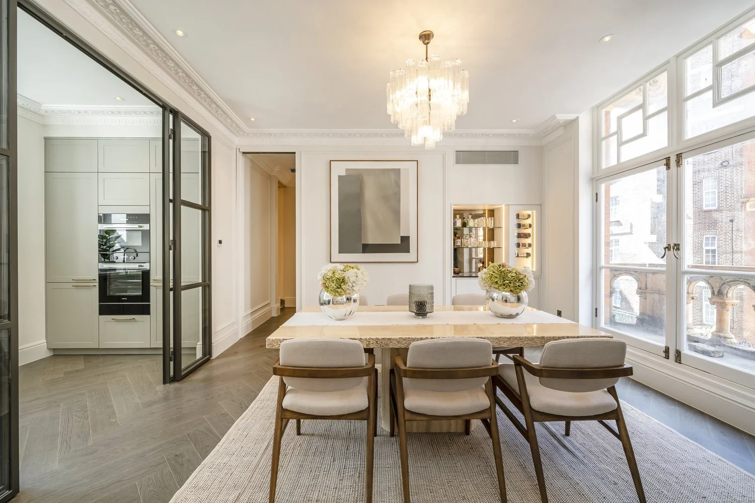 Bright, modern dining room with large windows, a chandelier, a marble dining table, six upholstered chairs, two vases with white flowers, and wall art.