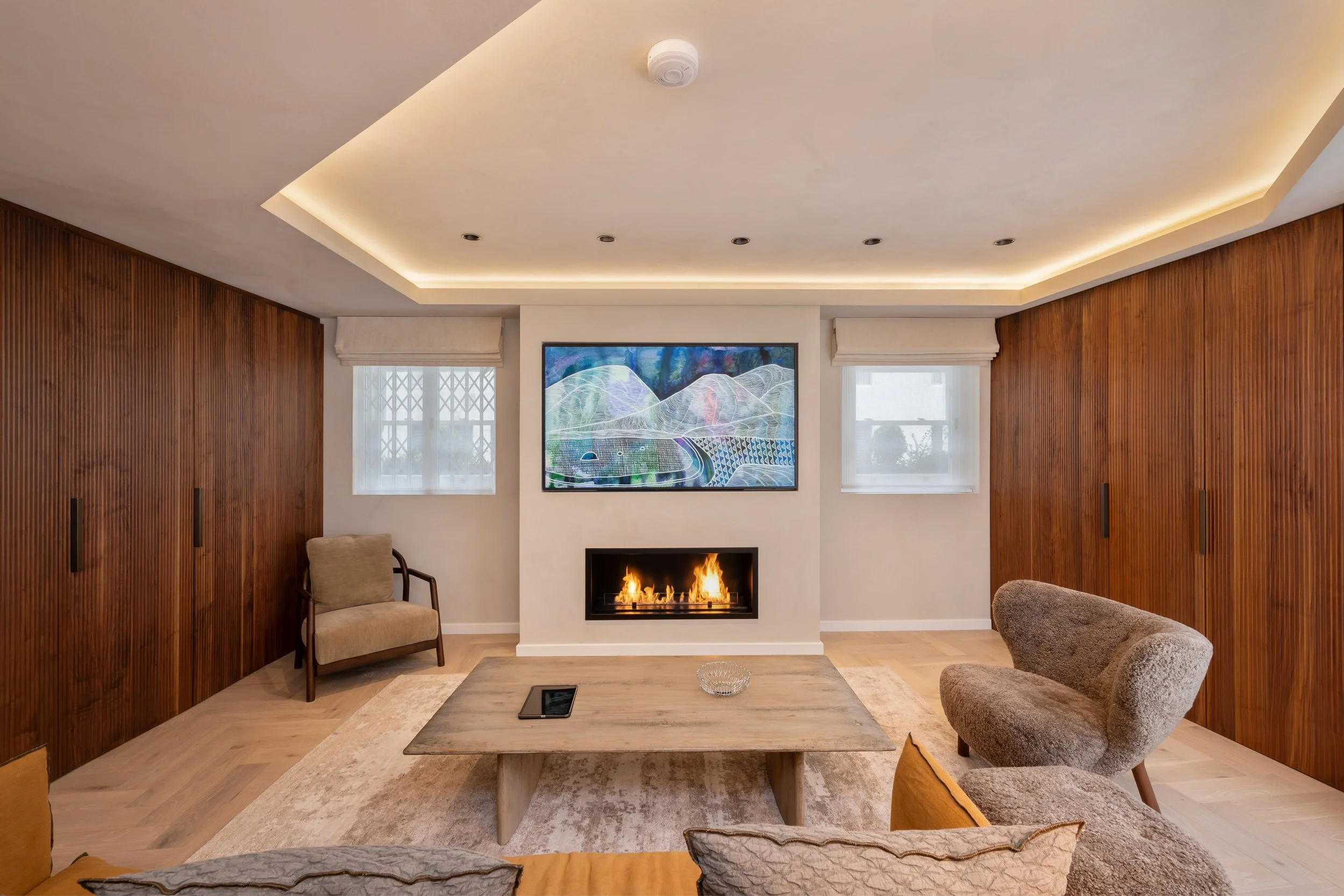 Living room with a modern fireplace, a flat-screen TV above it, a wooden coffee table, and comfortable chairs, with wood-paneled walls and two windows.