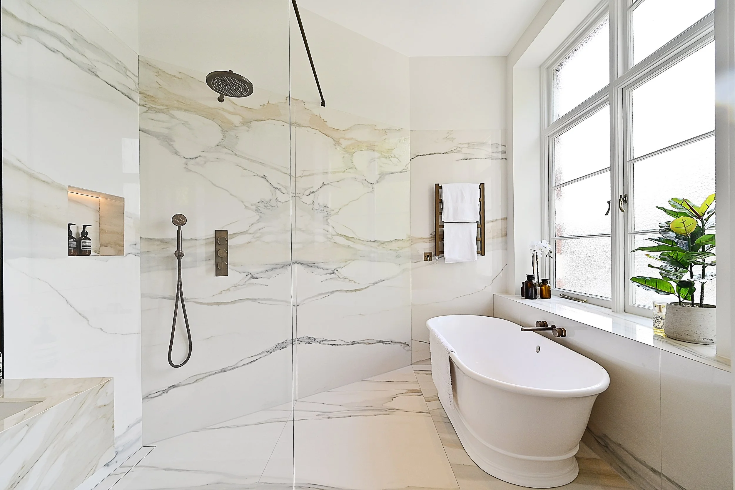 Modern bathroom with a freestanding bathtub, marble walls, large window with a window seat, and a walk-in shower with rainfall and handheld shower heads.