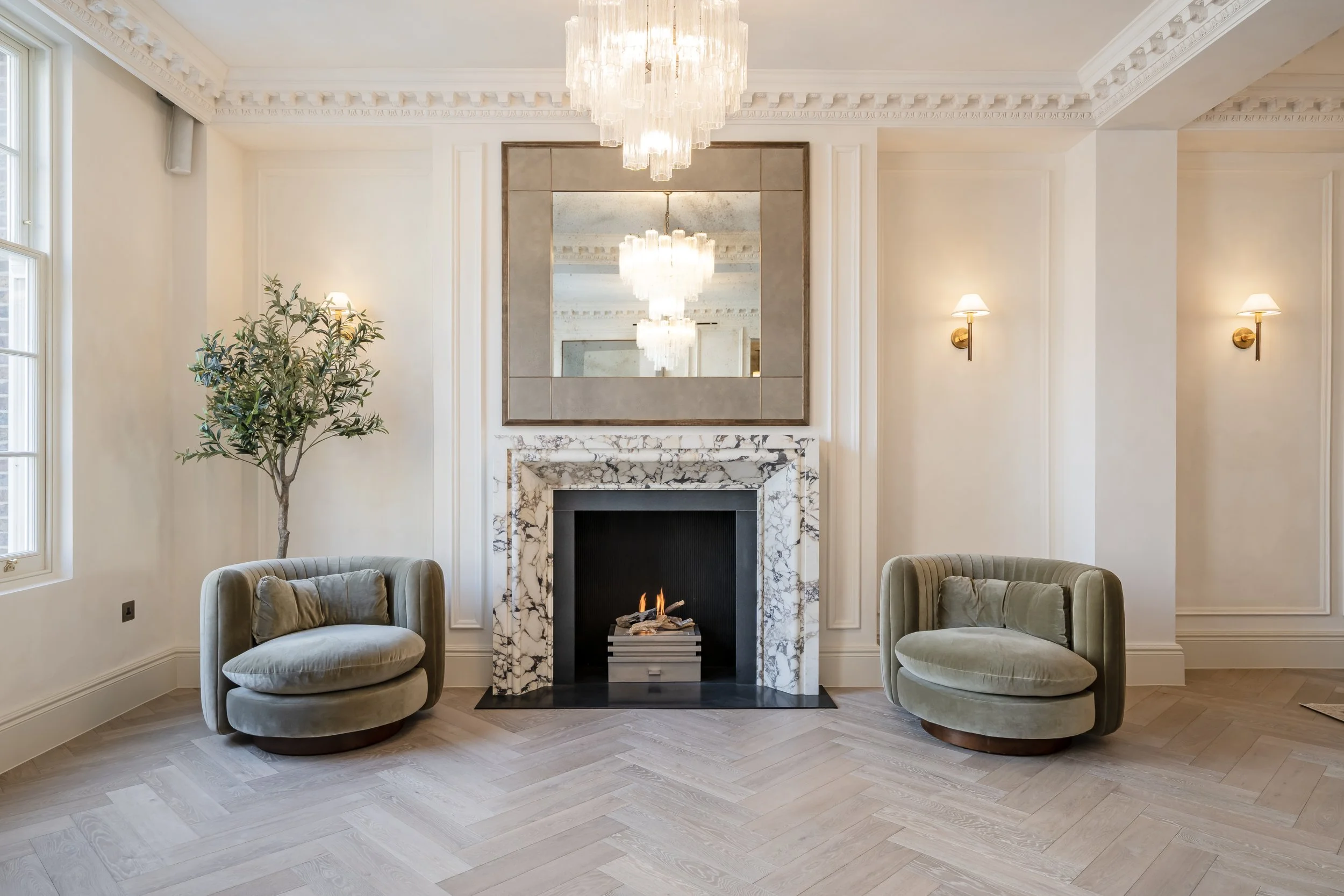 Elegant living room with a marble fireplace, large mirror, chandelier, two armchairs, wall sconces, and a potted tree.