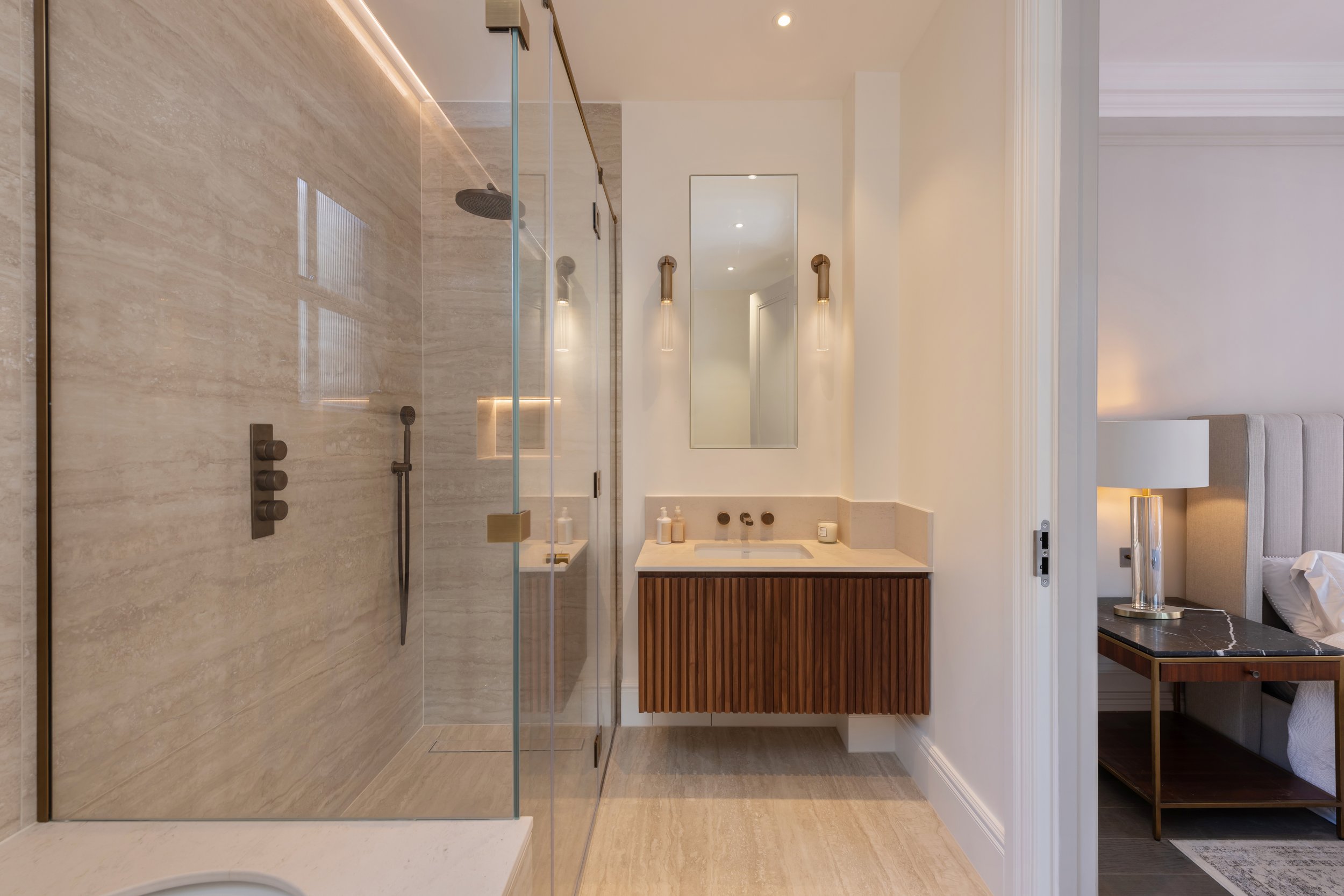 Modern bathroom with a glass shower enclosure, a white vanity with wooden slats, a large mirror, wall sconces, and a bedroom partially visible through an open door.