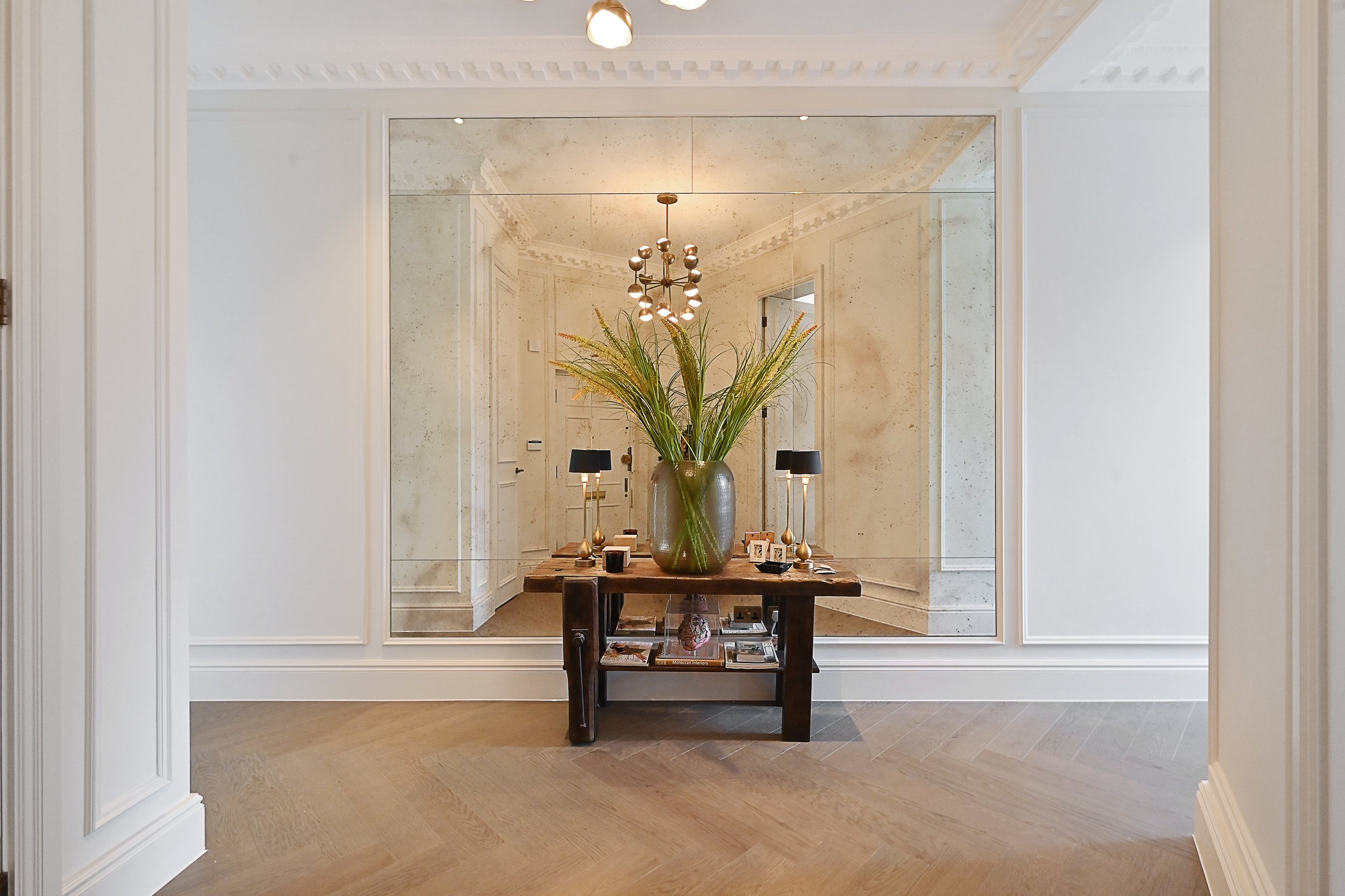 Elegant entryway with a large mirror, a wooden console table, a green vase with tall grass, two black lamps with gold bases, and a chandelier, with wooden flooring and cream walls.