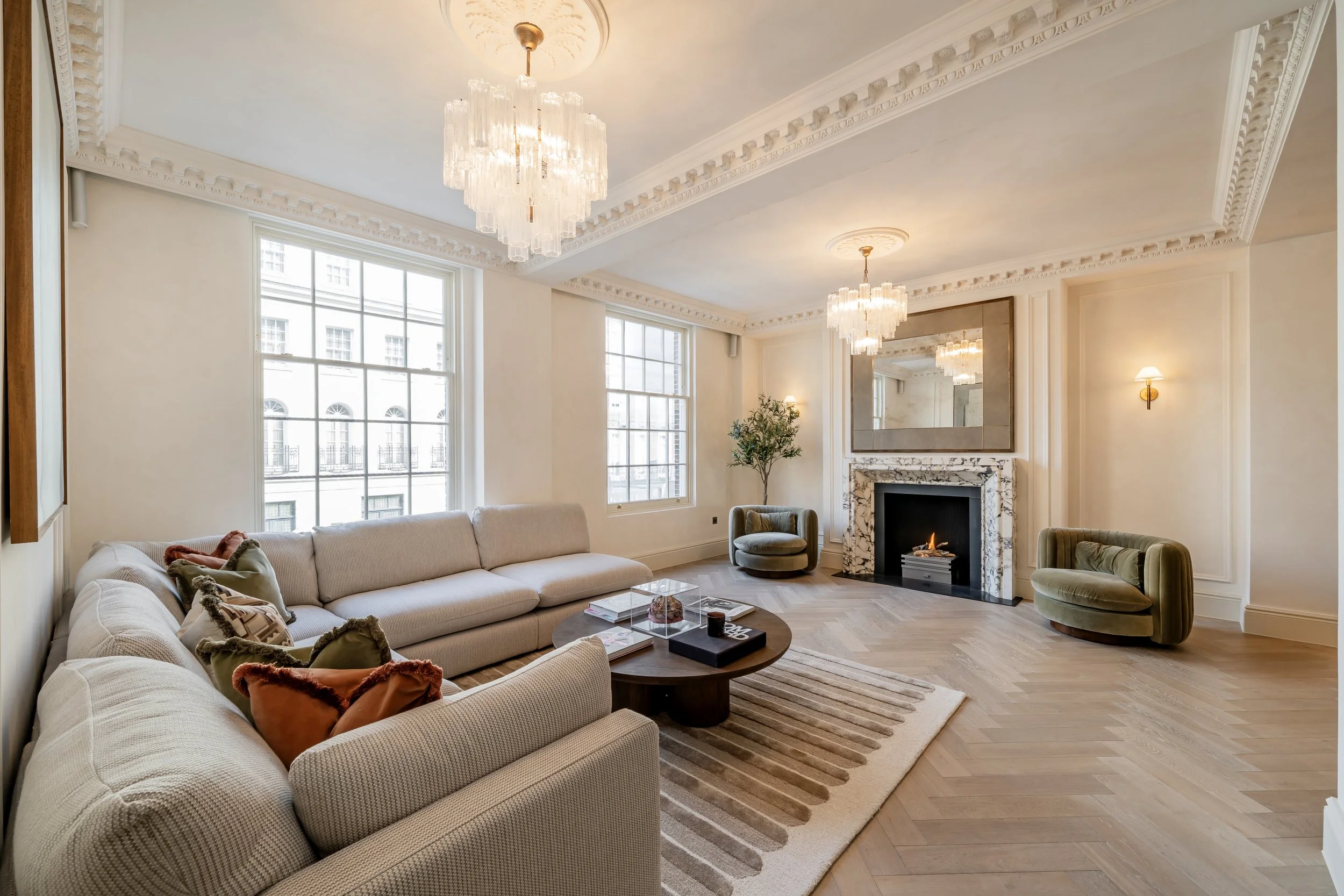 A spacious, elegantly decorated living room with large windows, a marble fireplace, a chandelier, and contemporary furniture including a long beige sofa, green chairs, and a round coffee table with books and decor.