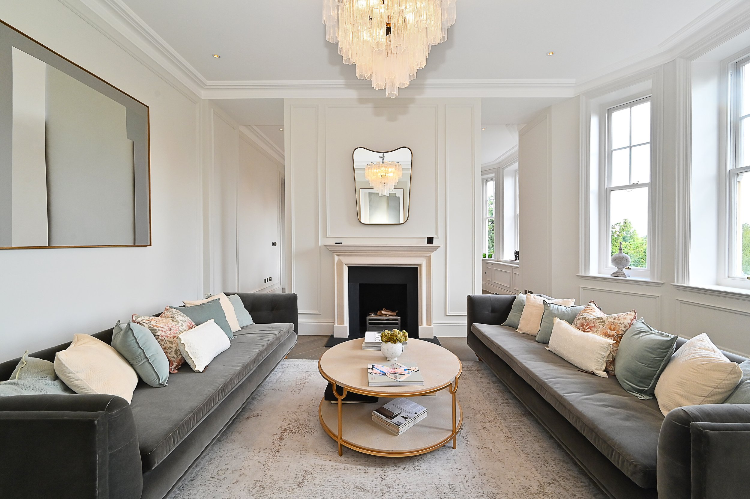 Elegant living room with white walls, a chandelier, a fireplace with a mirror above it, two long gray sofas with decorative pillows, a round coffee table with books and a small plant, and large windows letting in natural light.