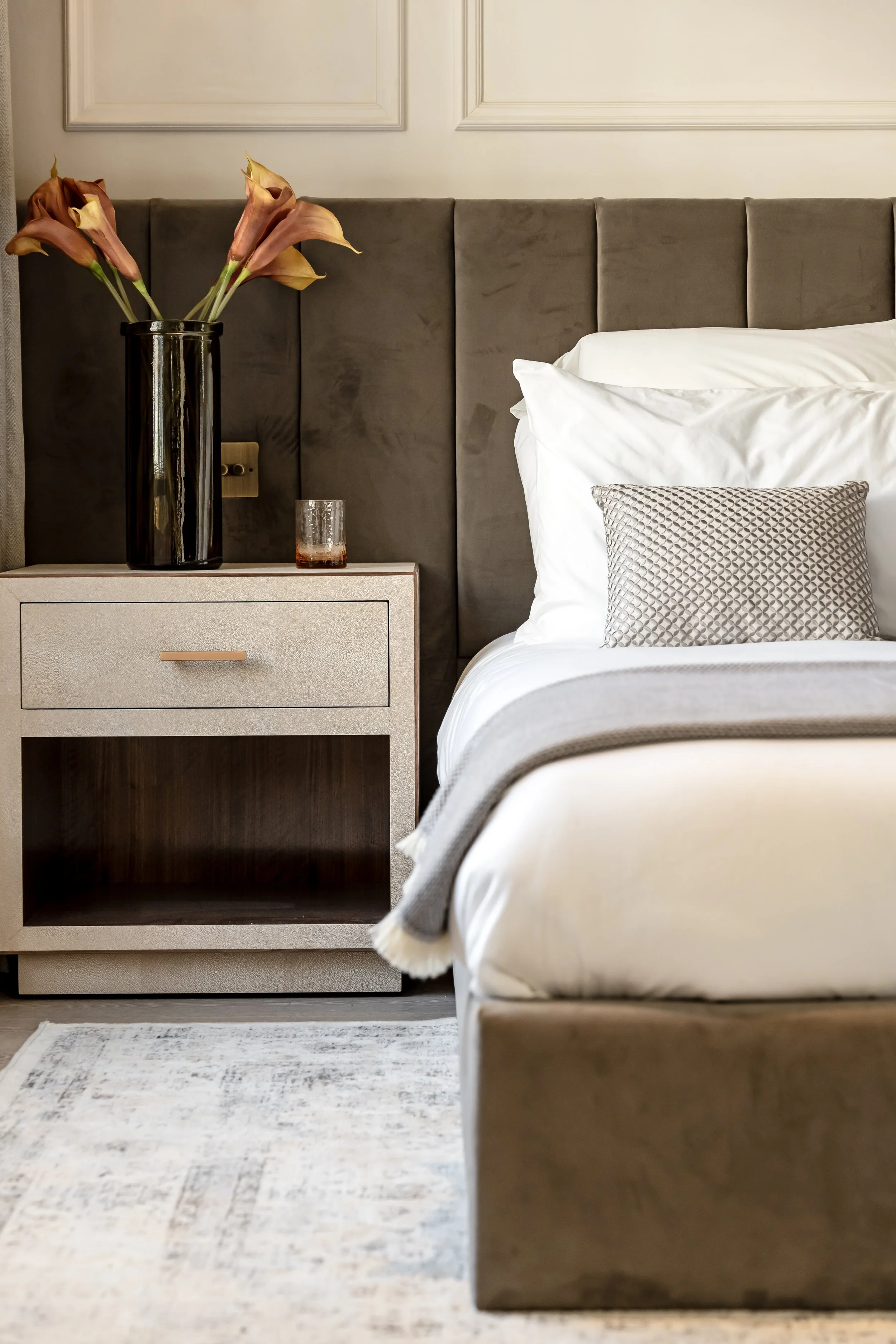 A bedroom with a bed, a beige nightstand, a black vase with pinkish-orange calla lilies, a glass with drink, and a patterned pillow.