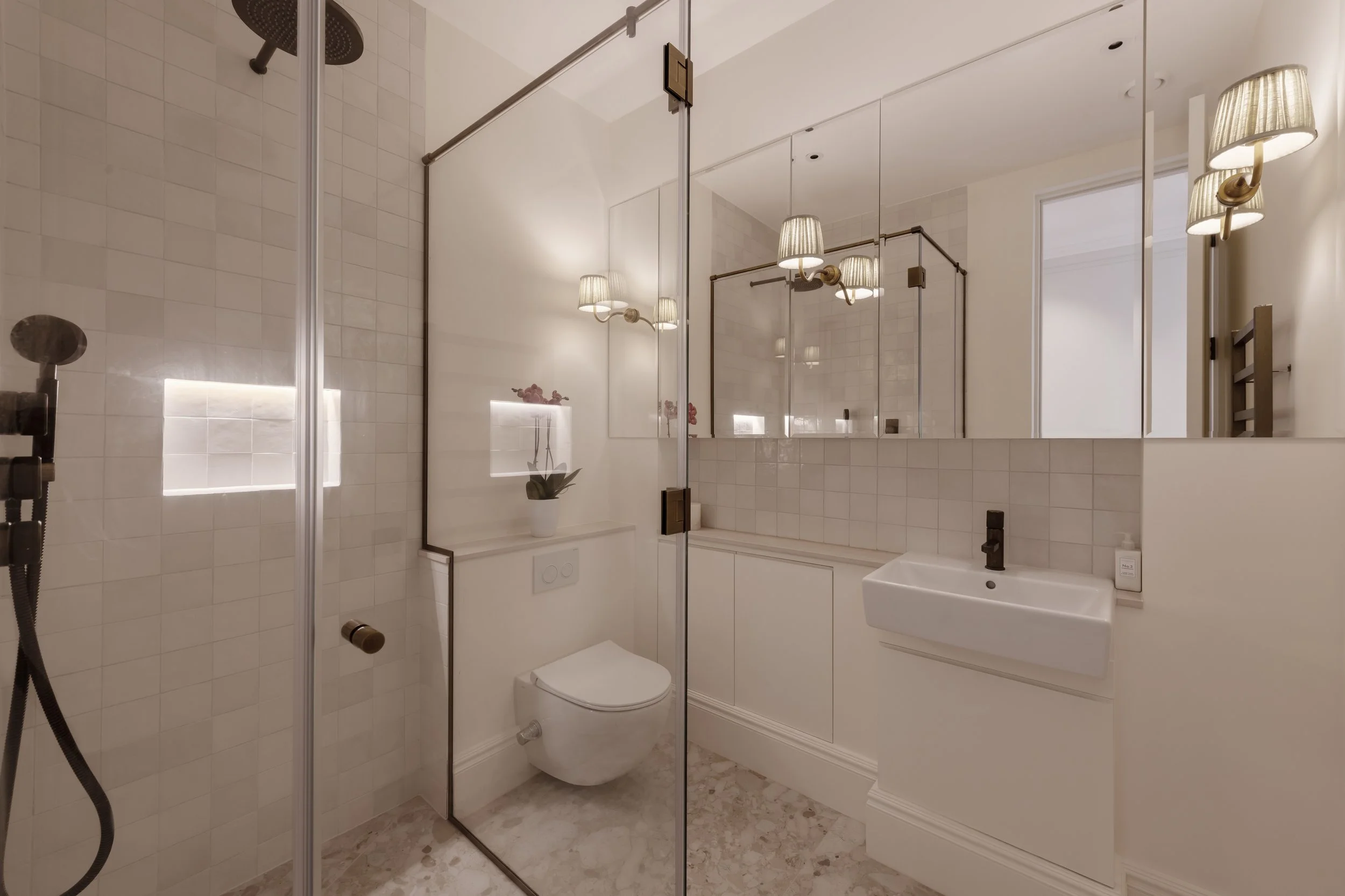 Modern bathroom with walk-in shower, wall-mounted toilet, small sink, large mirror, and warm lighting.