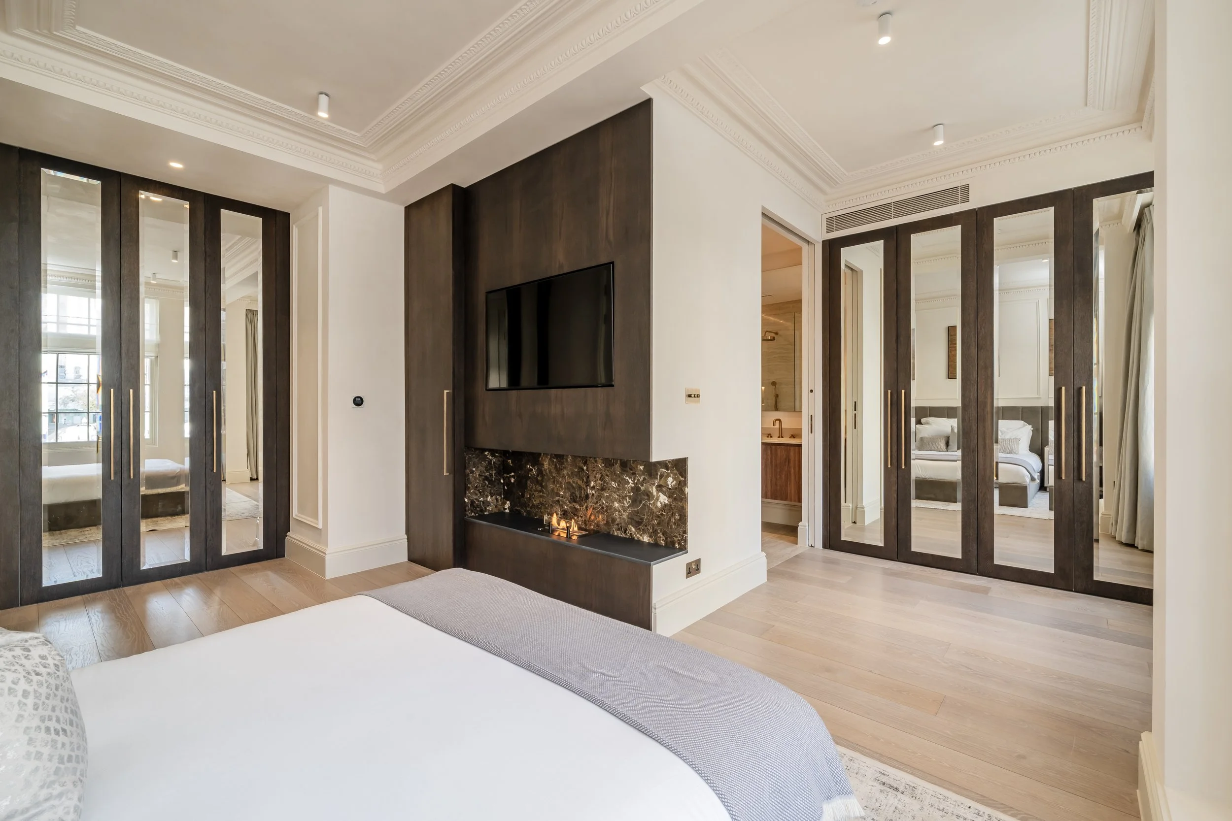Modern bedroom with a fireplace, flat-screen TV, mirrored closet doors, and a view into an adjacent bedroom and bathroom, with light wood flooring and elegant crown molding.