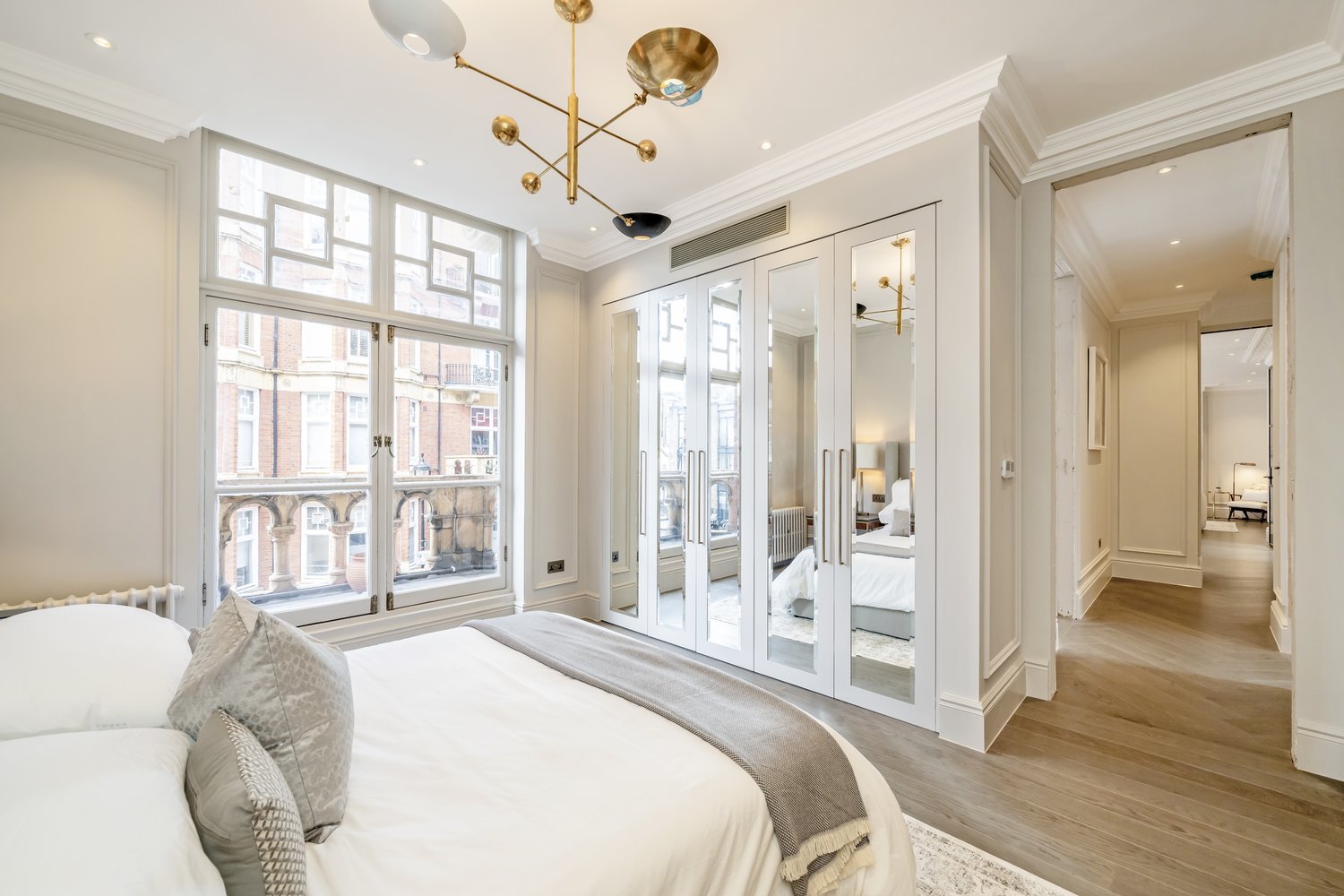 Bright bedroom with large windows, white walls, a bed with white bedding, decorative pillows, and a mirrored wardrobe with white doors, in a modern apartment.