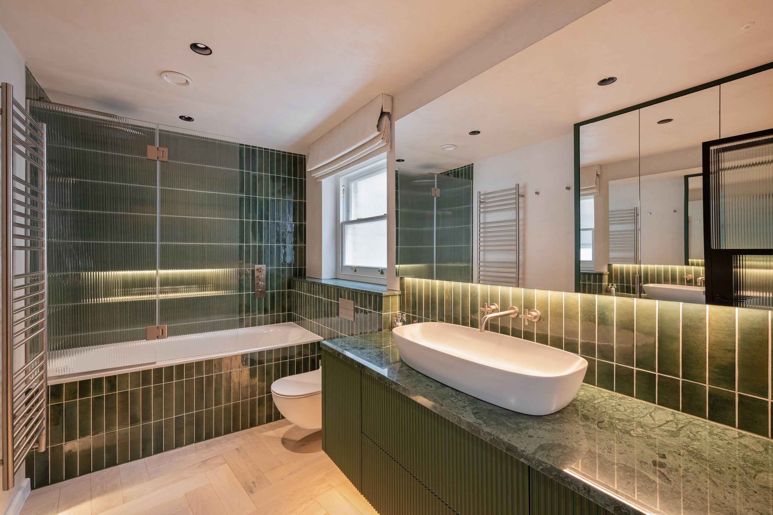 Modern bathroom with green tile walls, a white bathtub with a glass shower enclosure, a green marble countertop with a vessel sink, a large mirror, and wooden flooring.
