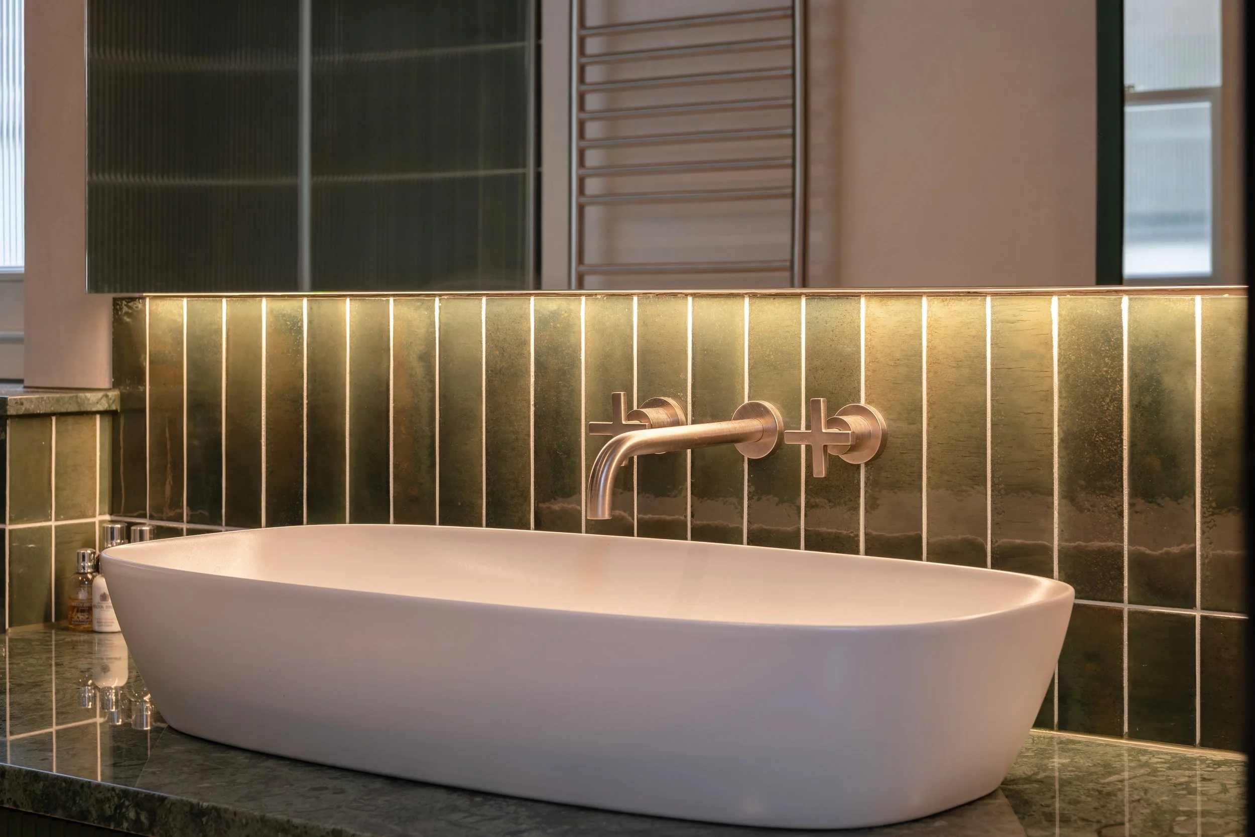 Modern bathroom with a white vessel sink, wall-mounted brushed metal faucet, green tile backsplash, and a large mirror reflecting the room’s interior.