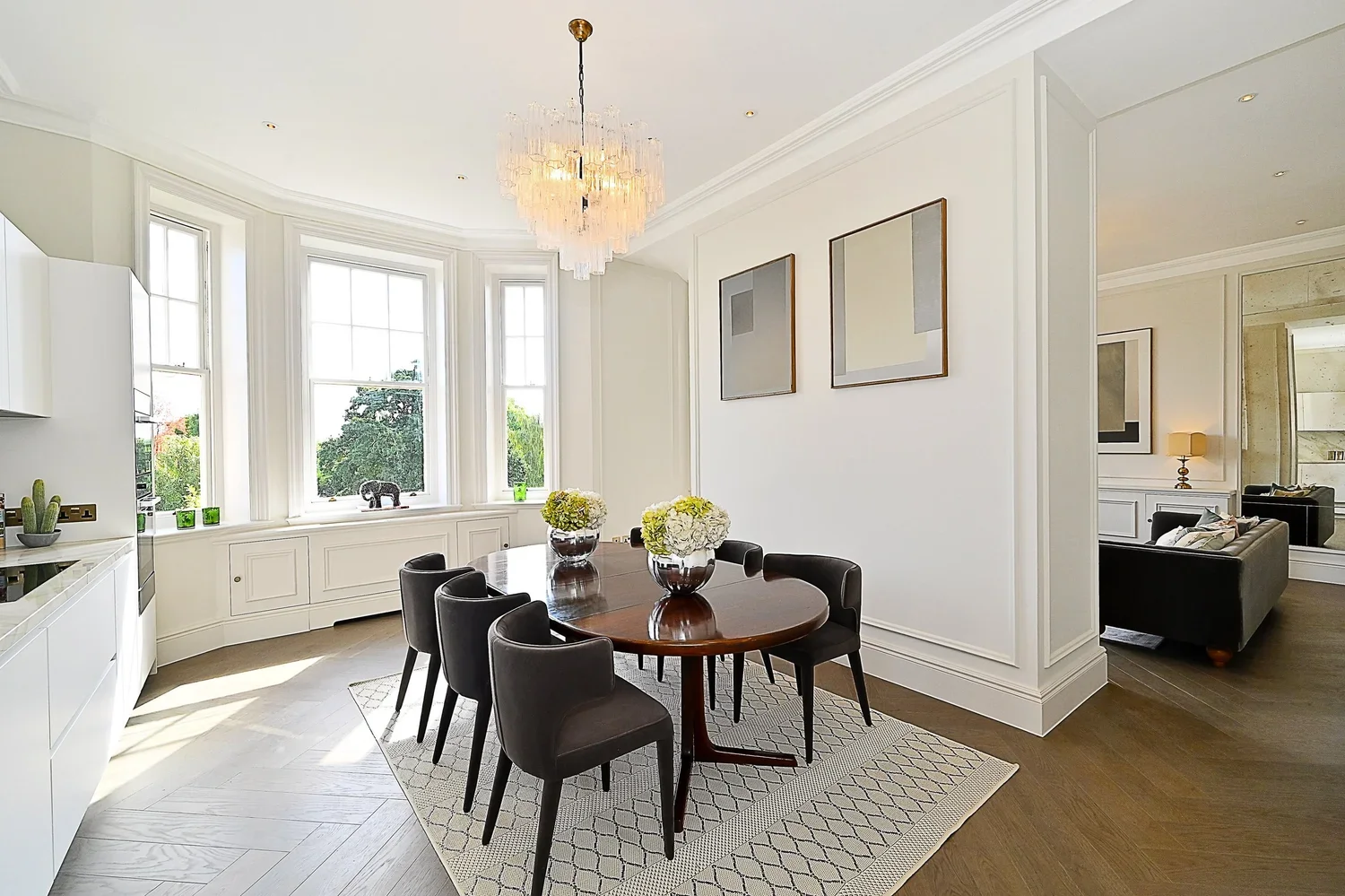 Bright dining room with a round wooden table, six black chairs, a chandelier, and three vases with white flowers, located by large windows with a view of greenery.