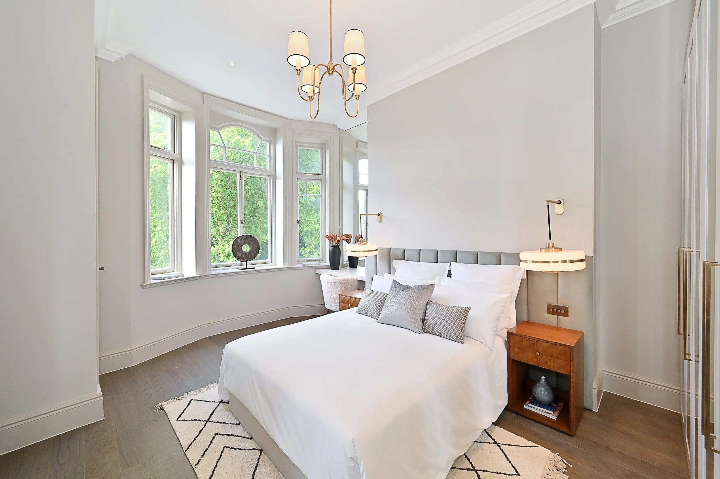 Bright bedroom with large bay window, white bedding, grey headboard, wooden nightstand, decorative vases, modern wall-mounted lamps, chandelier, and patterned rug.