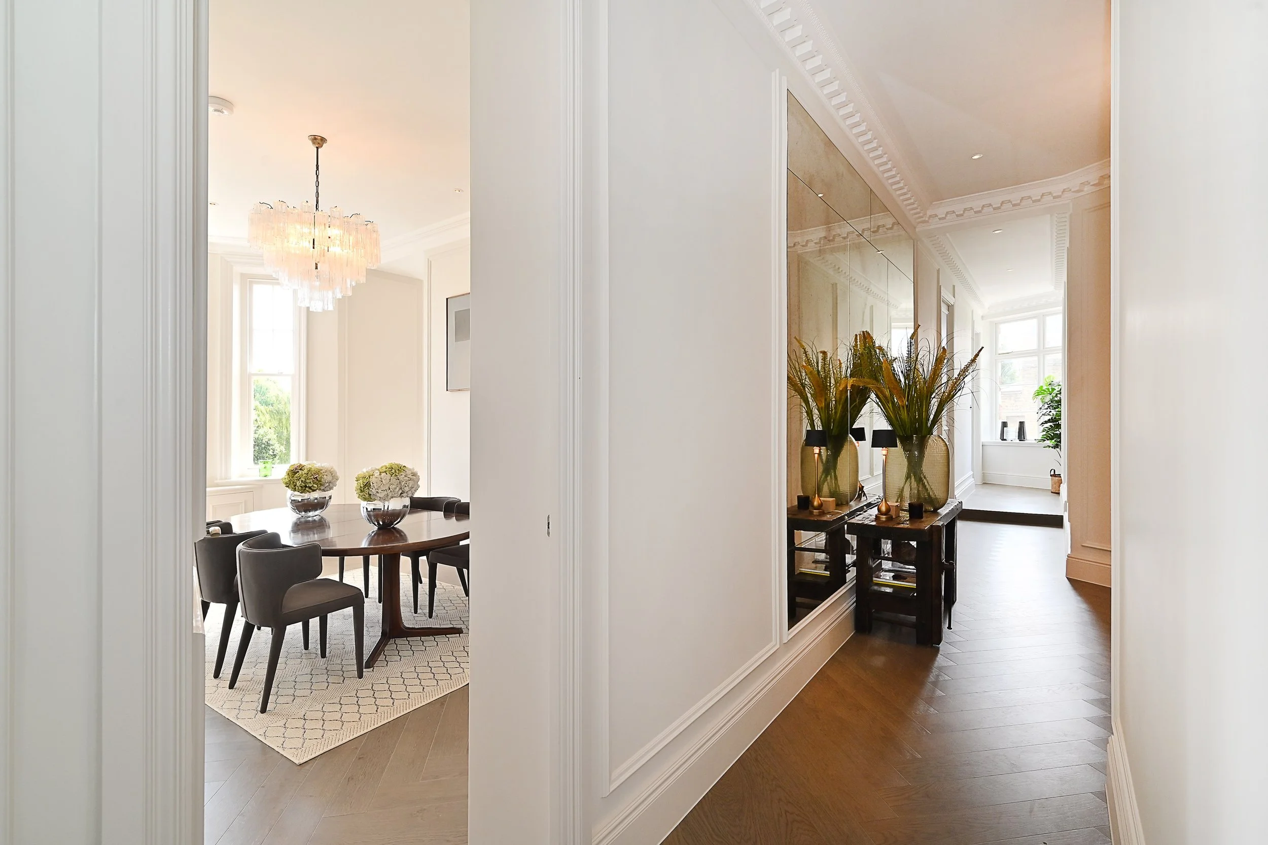 View of a bright, elegant hallway with white walls, crown molding, and wooden flooring leading to a dining room. The dining room has a round wooden table, surrounded by dark chairs, and a chandelier hanging overhead. Decorative floral arrangements ar