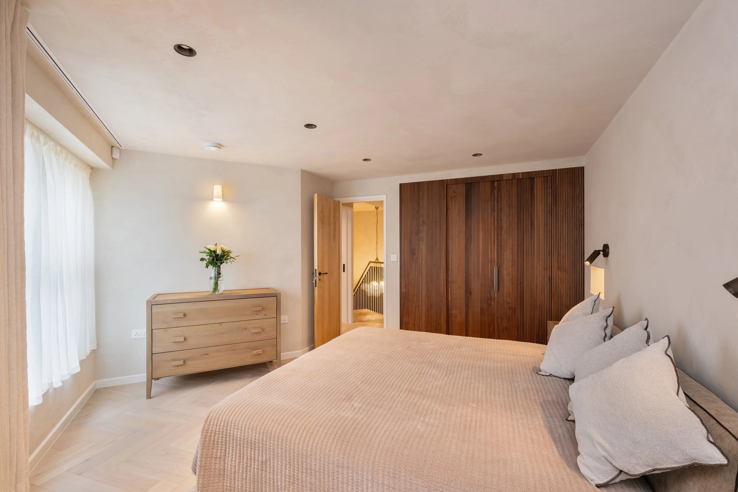 A bedroom featuring a bed with beige bedding and pillows, a wooden dresser with a vase of flowers, and a large wooden wardrobe. There is a door leading to a staircase and soft lighting.