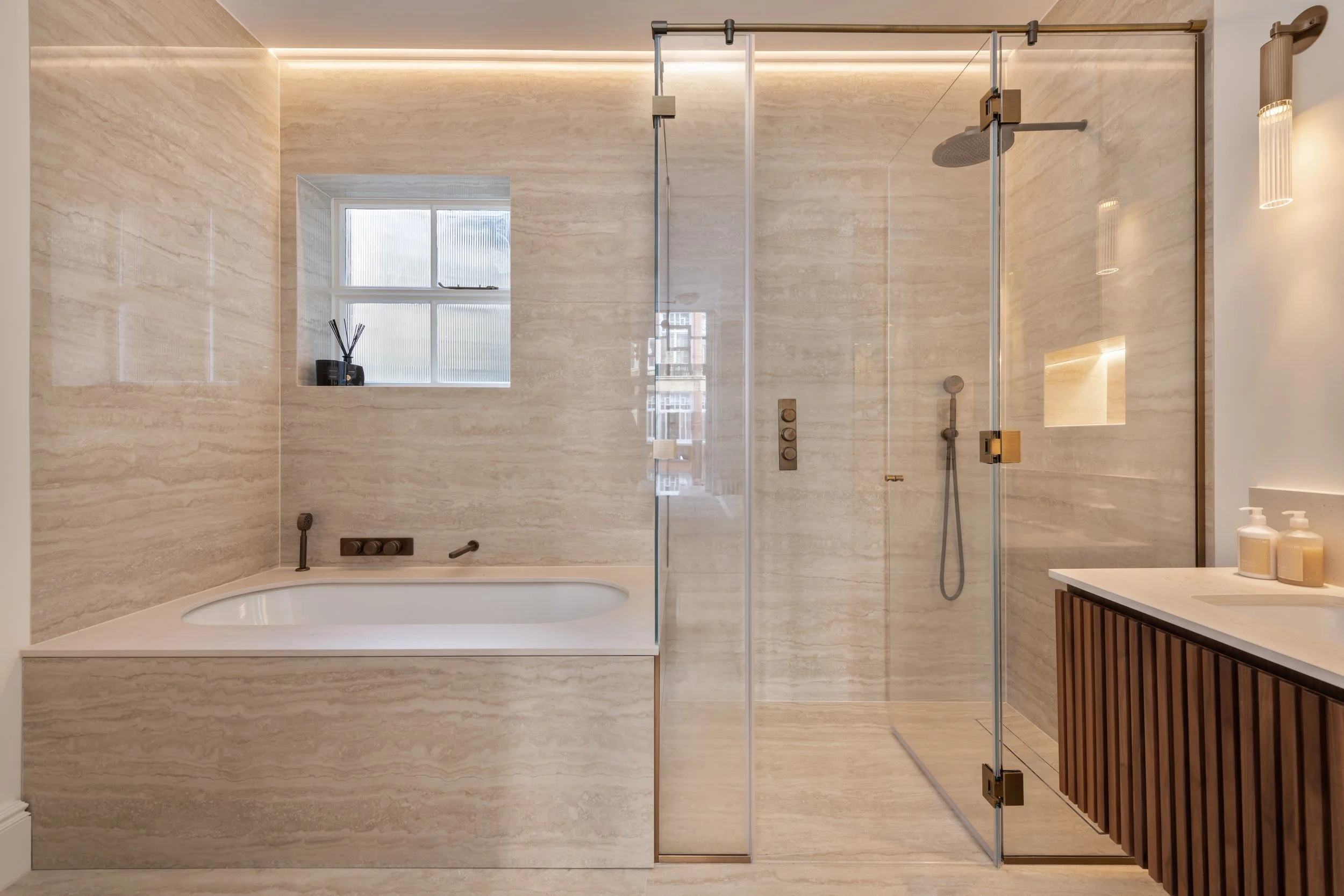 Modern bathroom with beige marble walls and floor, a built-in bathtub, glass-enclosed walk-in shower with brass fixtures, small window, wooden vanity with white countertop, and beige soap dispensers.